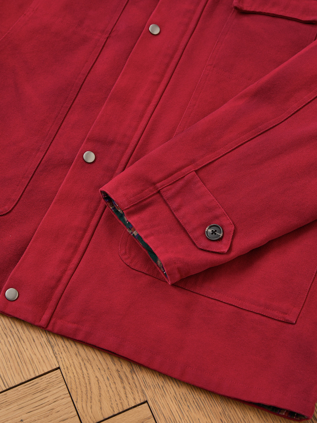 Close-up of the Found Canvas Utility Barn Jacket in red, featuring silver snap buttons, a corduroy collar, and a buttoned cuff, displayed on a wooden floor.