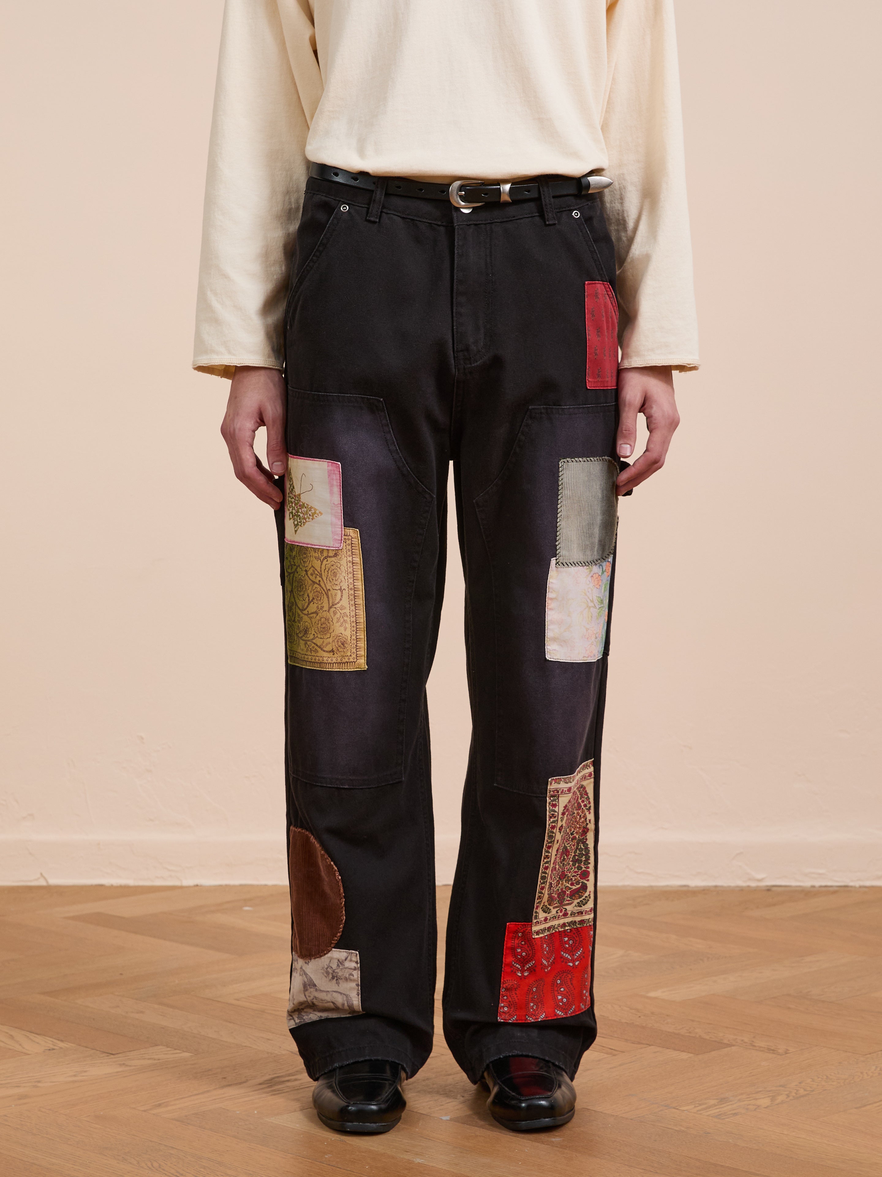 A person wears FOUND's Tapestry Carpenter Canvas Pants—unisex, relaxed fit, black with colorful Pakistani quilt-inspired patchwork—paired with a cream long sleeve shirt, black belt and shoes, standing on a wooden floor against a beige wall.