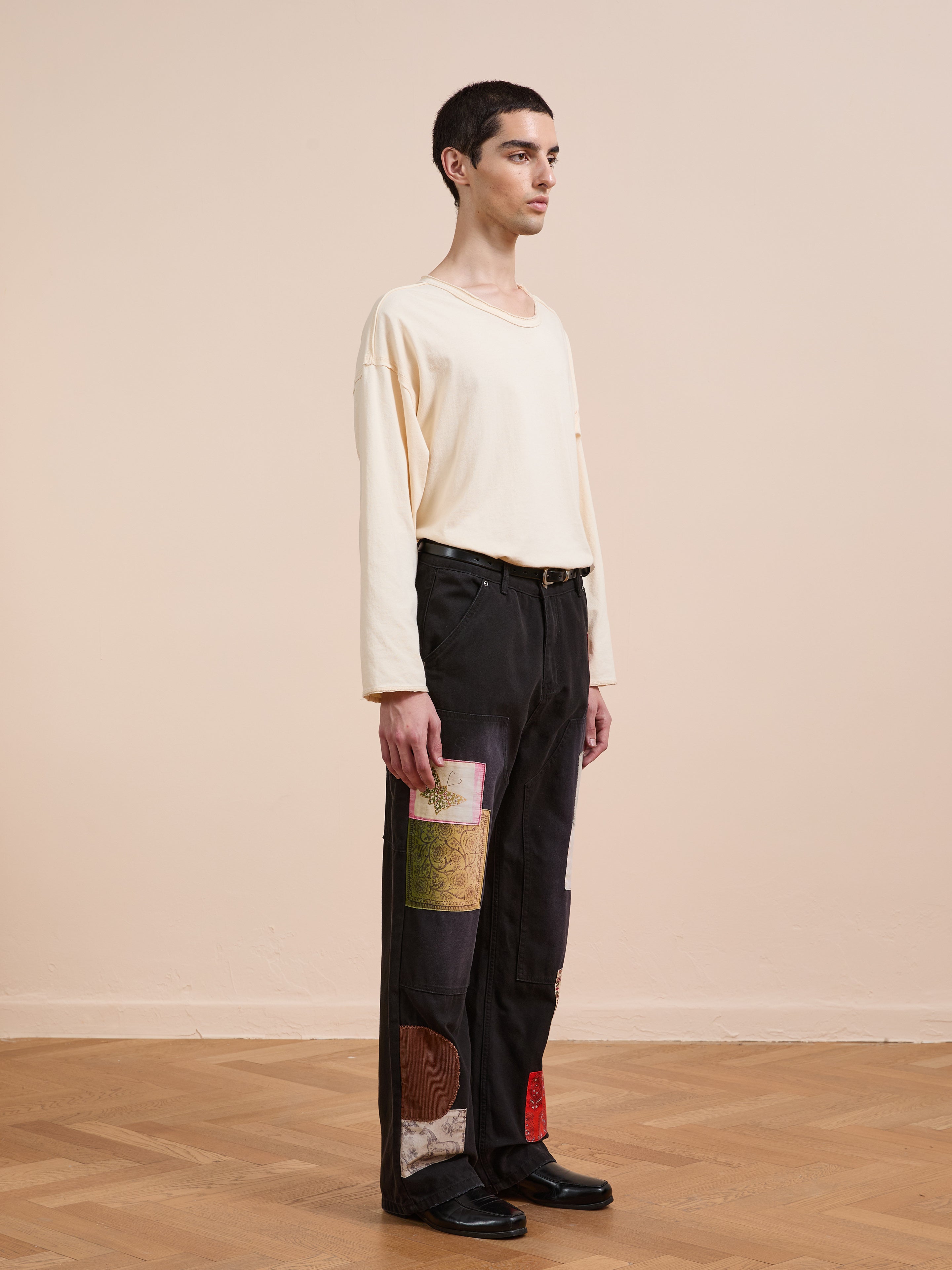 Indoors on a wooden floor, a person faces left in profile, wearing a cream long-sleeve shirt and FOUND Tapestry Carpenter Canvas Pants—unisex black pants featuring colorful patchwork inspired by Pakistani quilts.