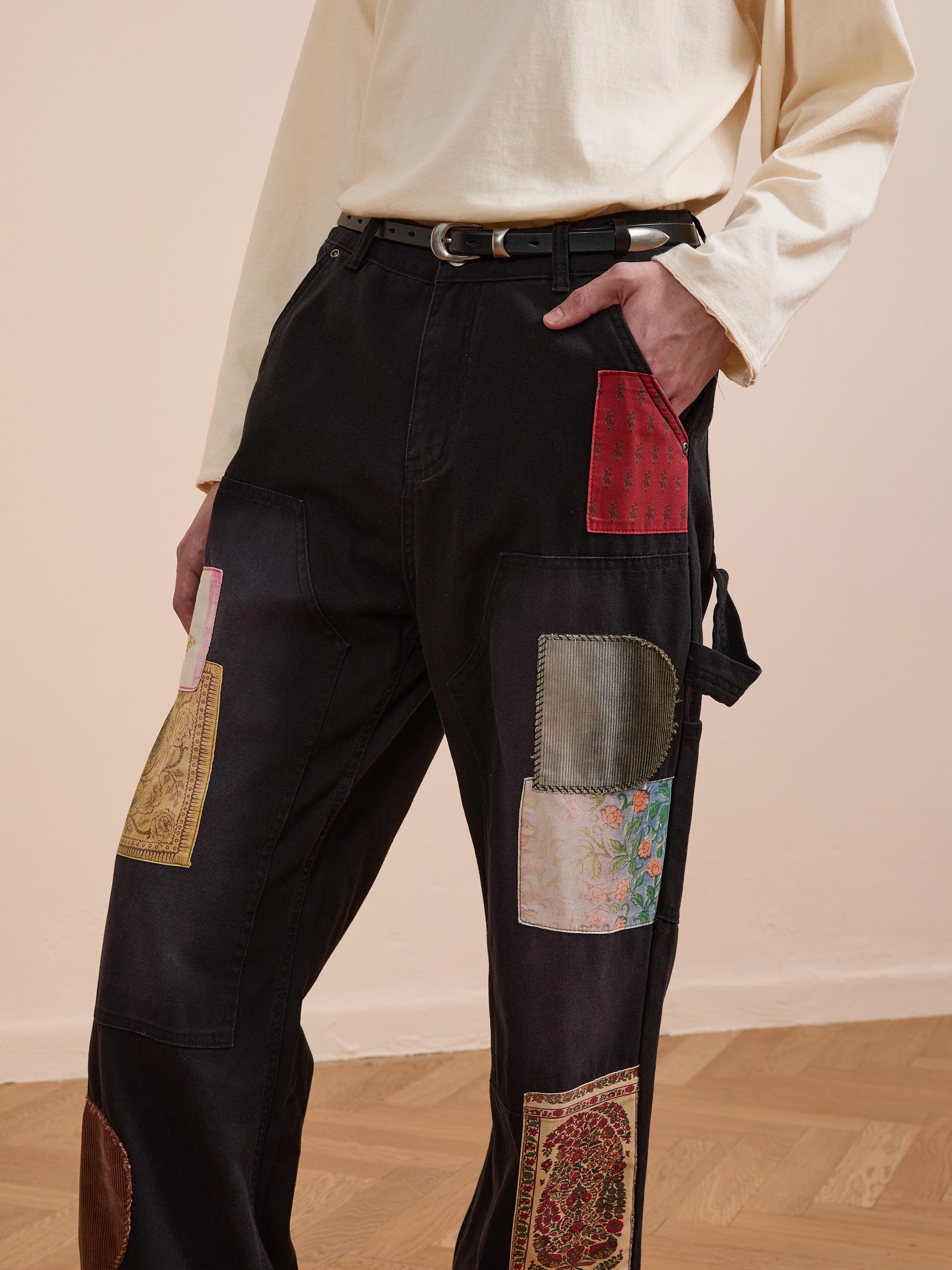 A person models FOUND's Tapestry Carpenter Canvas Pants—unisex, relaxed fit black pants featuring colorful Pakistani quilt-inspired patchwork—paired with a beige long-sleeve shirt, standing on a wooden floor, hand in pocket.