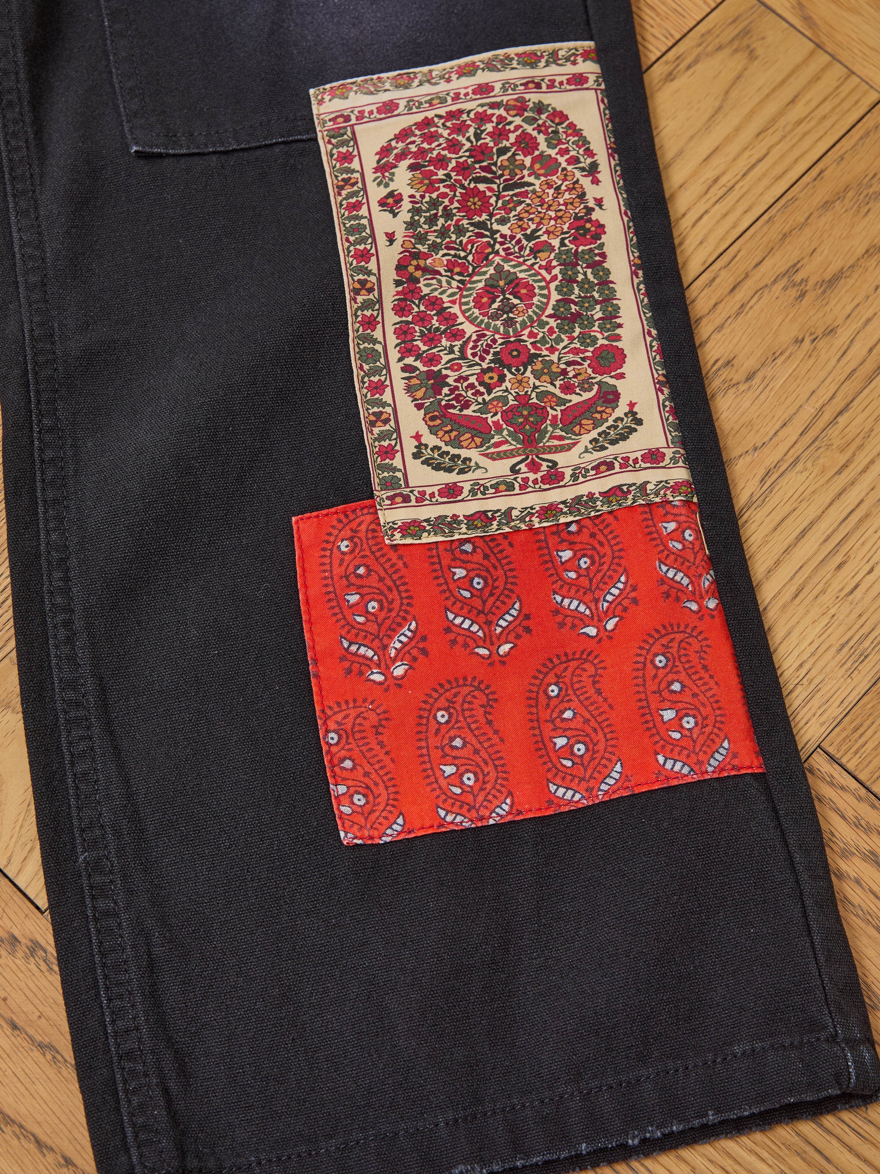 A close-up of FOUND’s Tapestry Carpenter Canvas Pants shows black fabric with sewn floral and red paisley patches, displayed on a wooden floor.