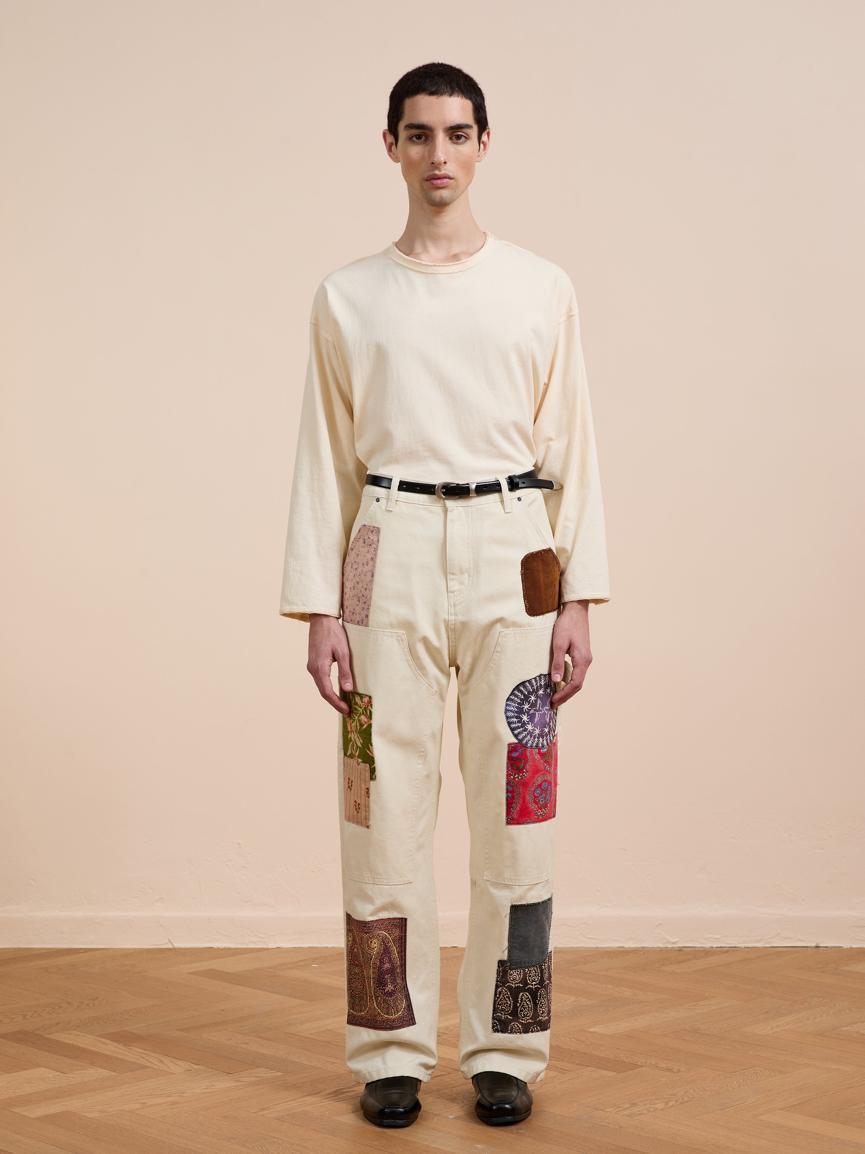 A person stands indoors against a plain beige background, wearing a cream long-sleeve shirt and FOUND’s Tapestry Carpenter Canvas Pants with antique Pakistani quilt-inspired print, paired with black shoes and a black belt.