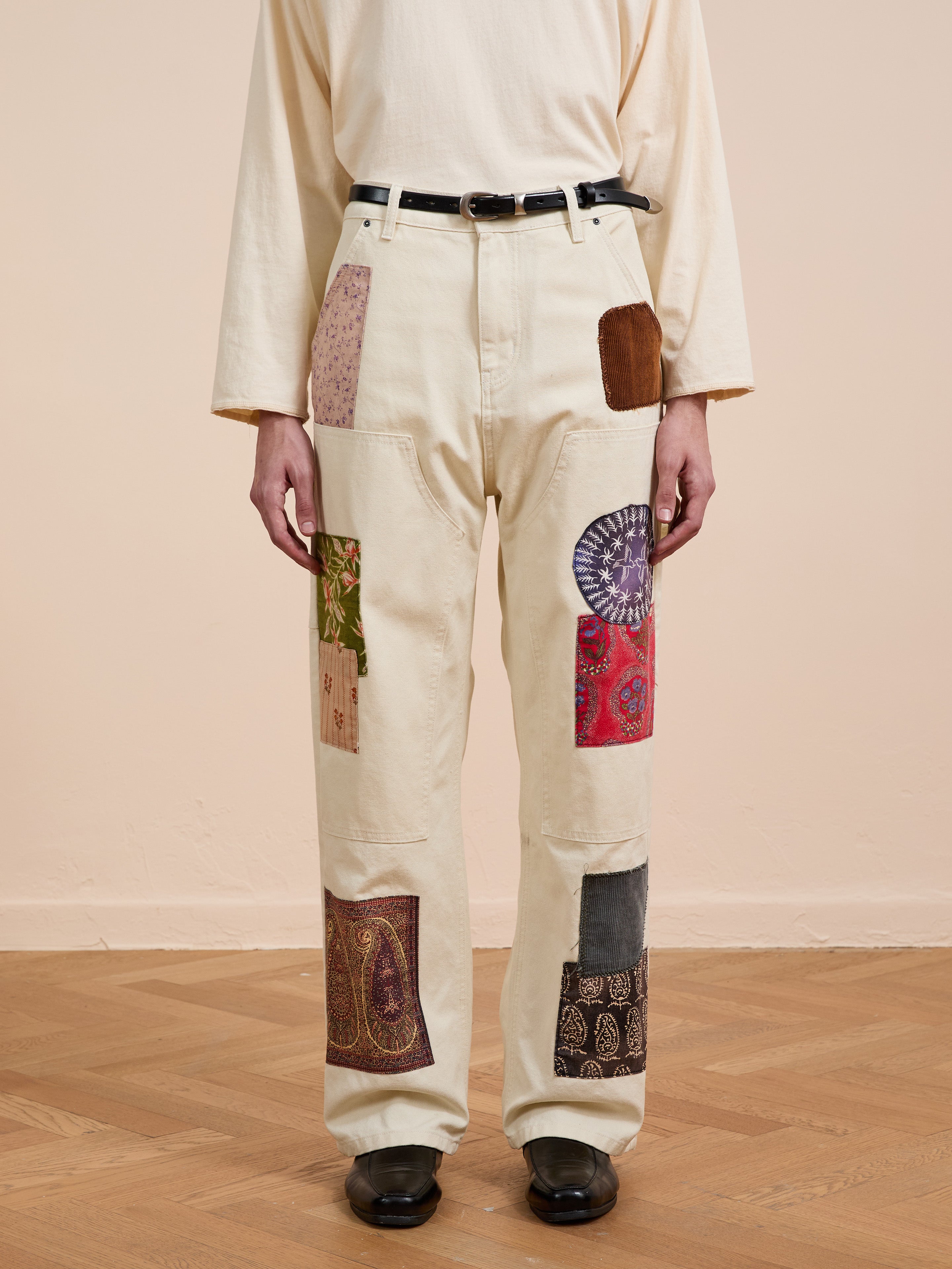 A person wears FOUND's Tapestry Carpenter Canvas Pants styled with a cream long-sleeve shirt, black belt, and shoes on a wooden floor—capturing vintage upholstery-inspired fashion.
