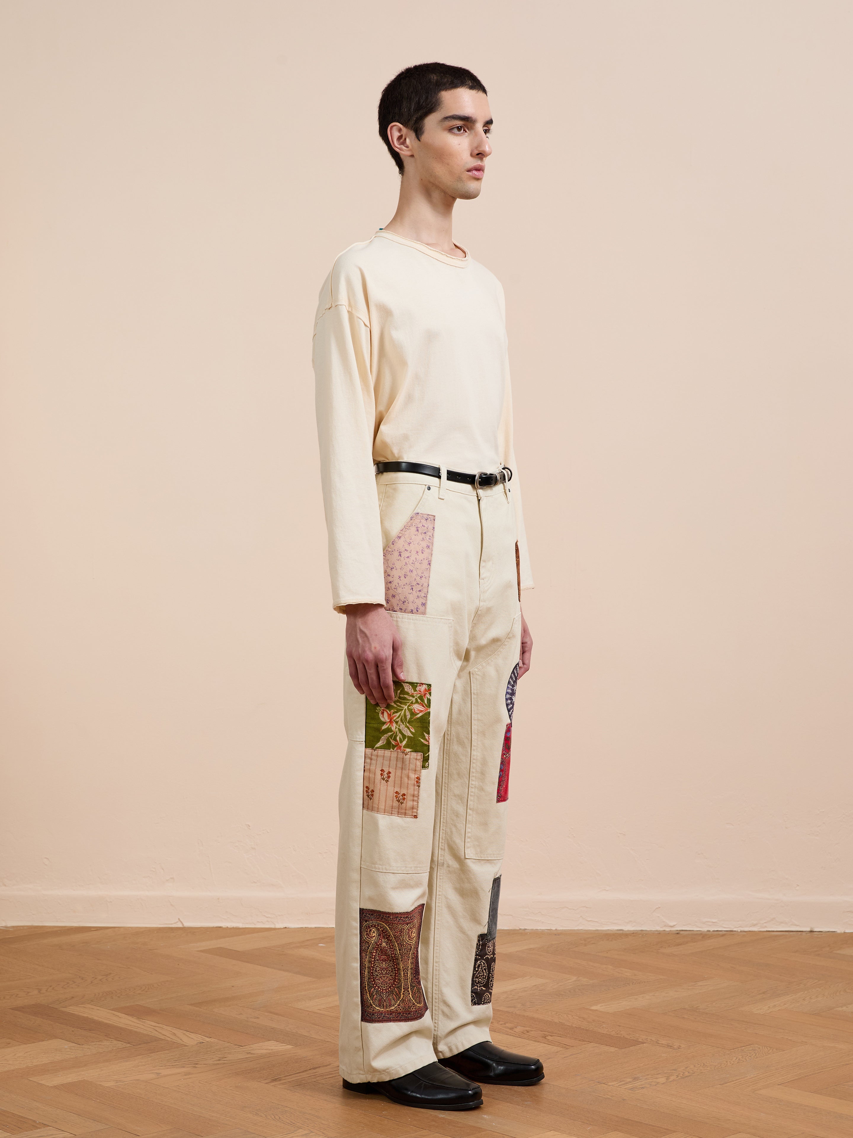 Indoors, a person wears a cream long-sleeve shirt and FOUND’s Tapestry Carpenter Canvas Pants, which feature vintage upholstery-inspired printed twill patchwork, styled with black shoes and a black belt.