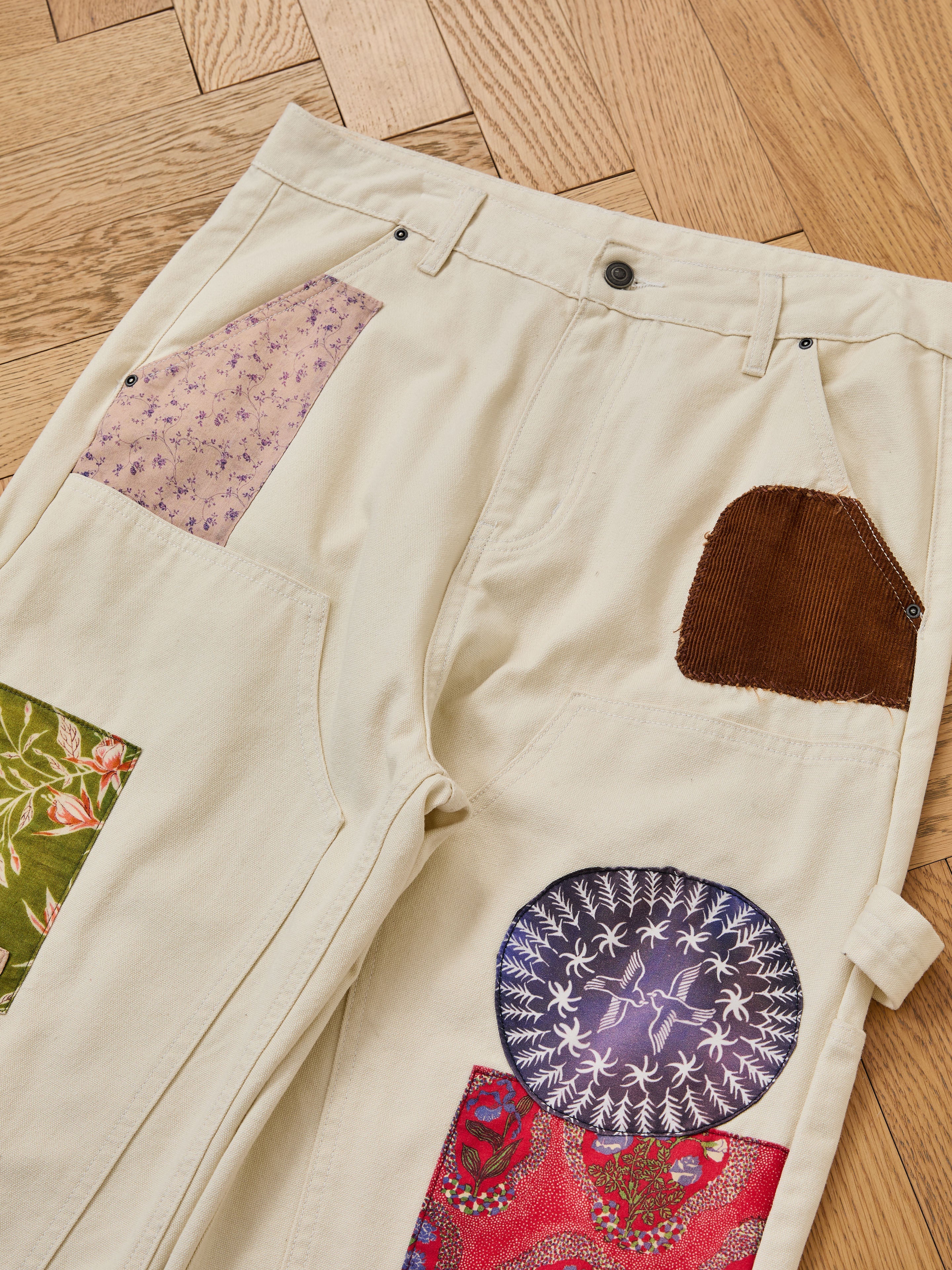 The FOUND Tapestry Carpenter Canvas Pants are unisex, relaxed-fit pants featuring colorful patchwork with floral, corduroy, and paisley fabrics, inspired by Pakistani quilt traditions. Shown laid flat on a wooden parquet floor.