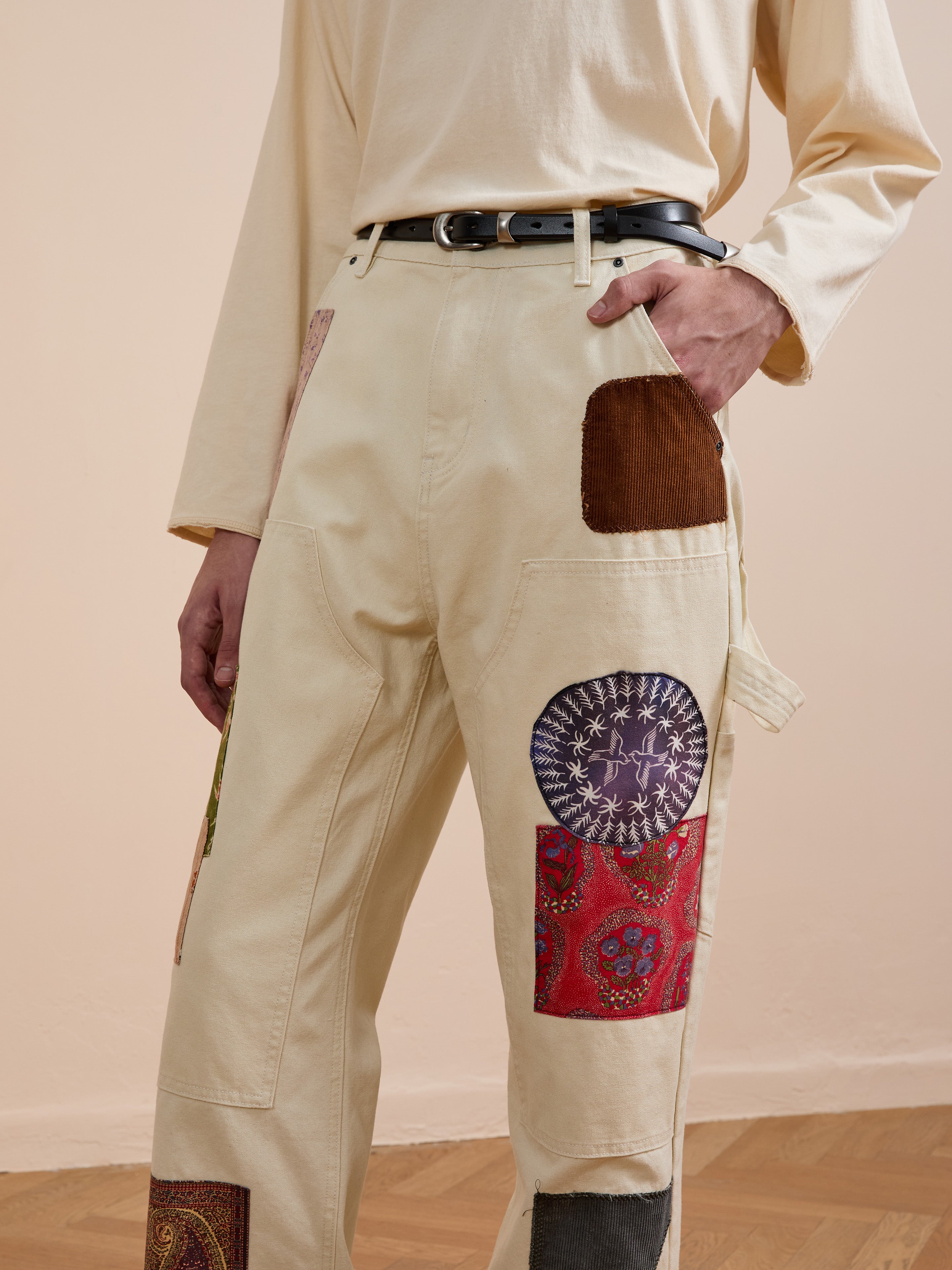 A person wears FOUND Tapestry Carpenter Canvas Pants featuring Pakistani quilt-inspired patchwork, paired with a black belt and cream unisex relaxed-fit long-sleeve shirt, standing indoors on a wooden floor.