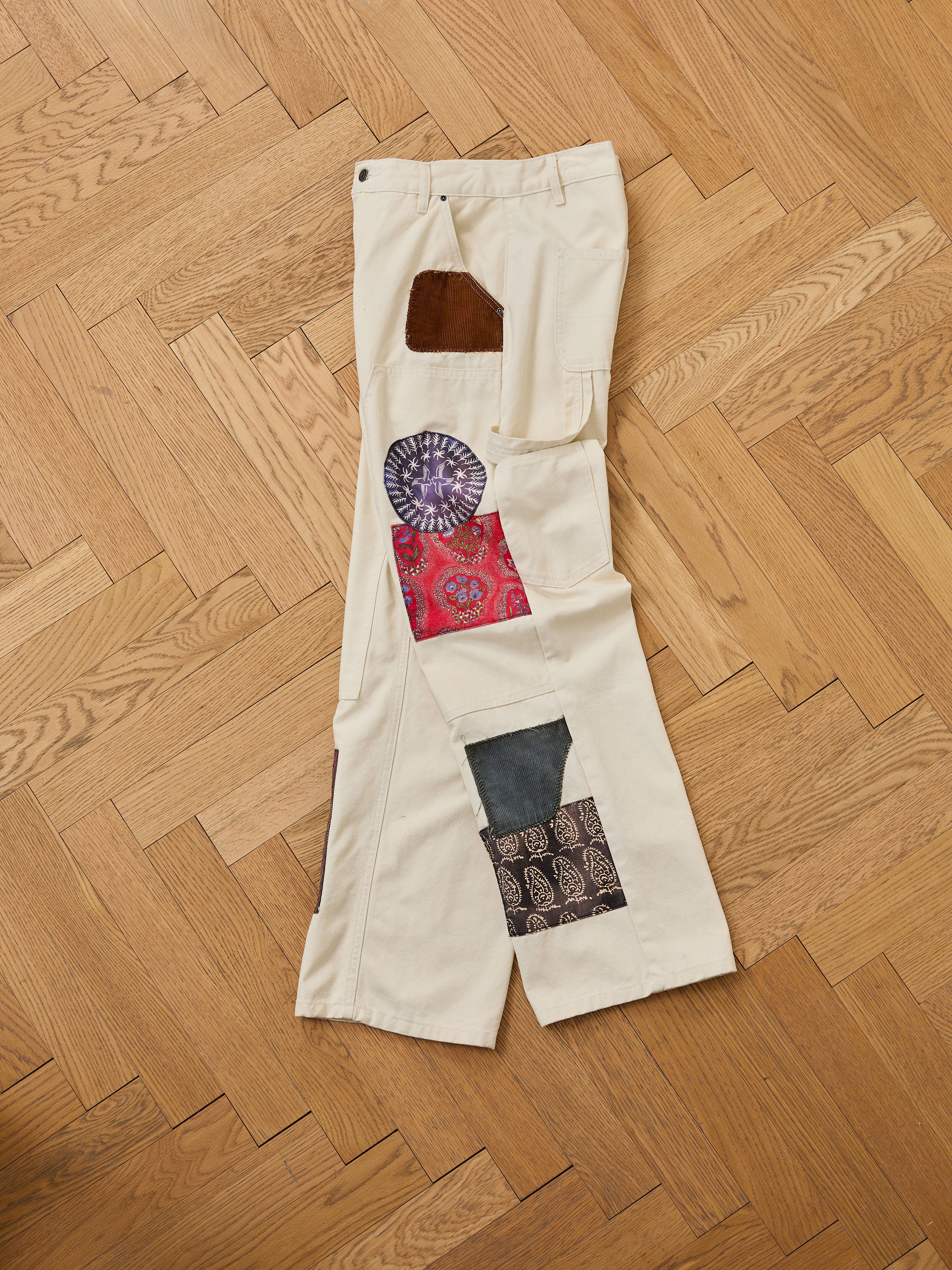The FOUND Tapestry Carpenter Canvas Pants are cream-colored and made of printed twill, displayed flat on a wooden floor with multicolored fabric patches sewn onto the legs.