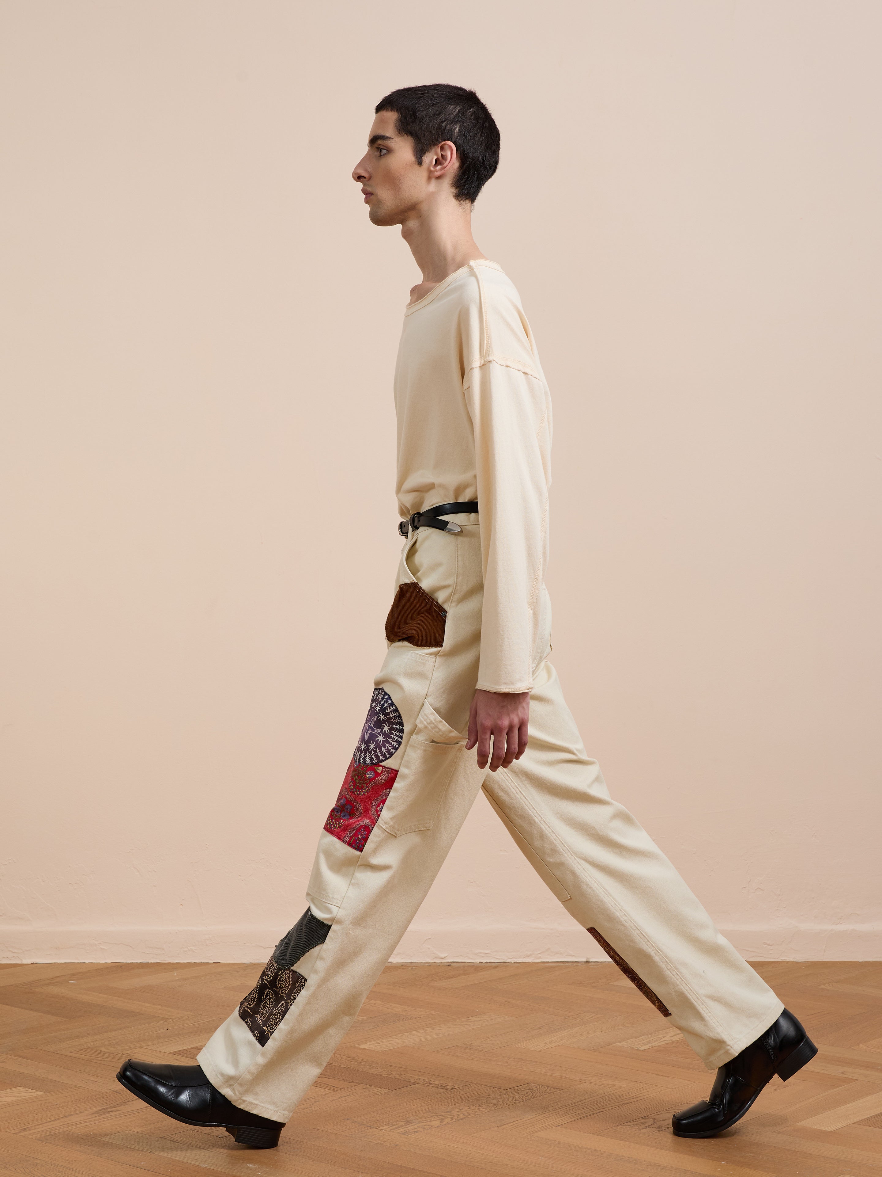 A person wearing FOUND's Tapestry Carpenter Canvas Pants walks across a wooden floor in cream-colored clothing and black shoes, set against a plain beige wall.