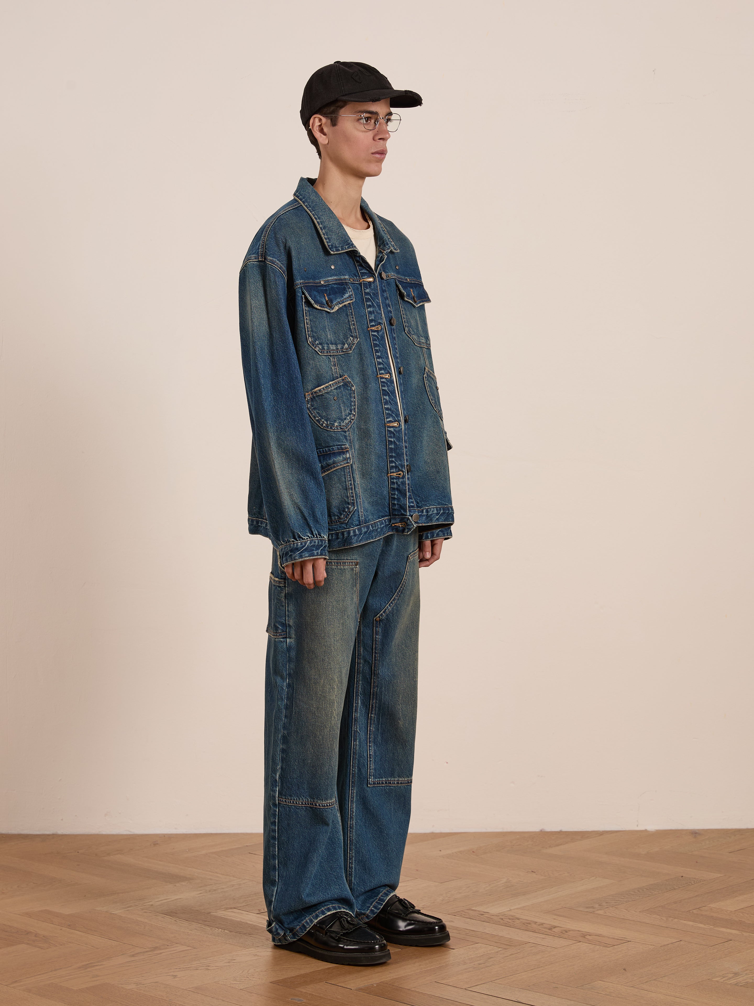 Wearing a Denim Carpenter Jacket by FOUND, this individual in jeans, glasses, and a black cap stands on a wooden floor against a beige backdrop, showcasing the timeless appeal of workwear.