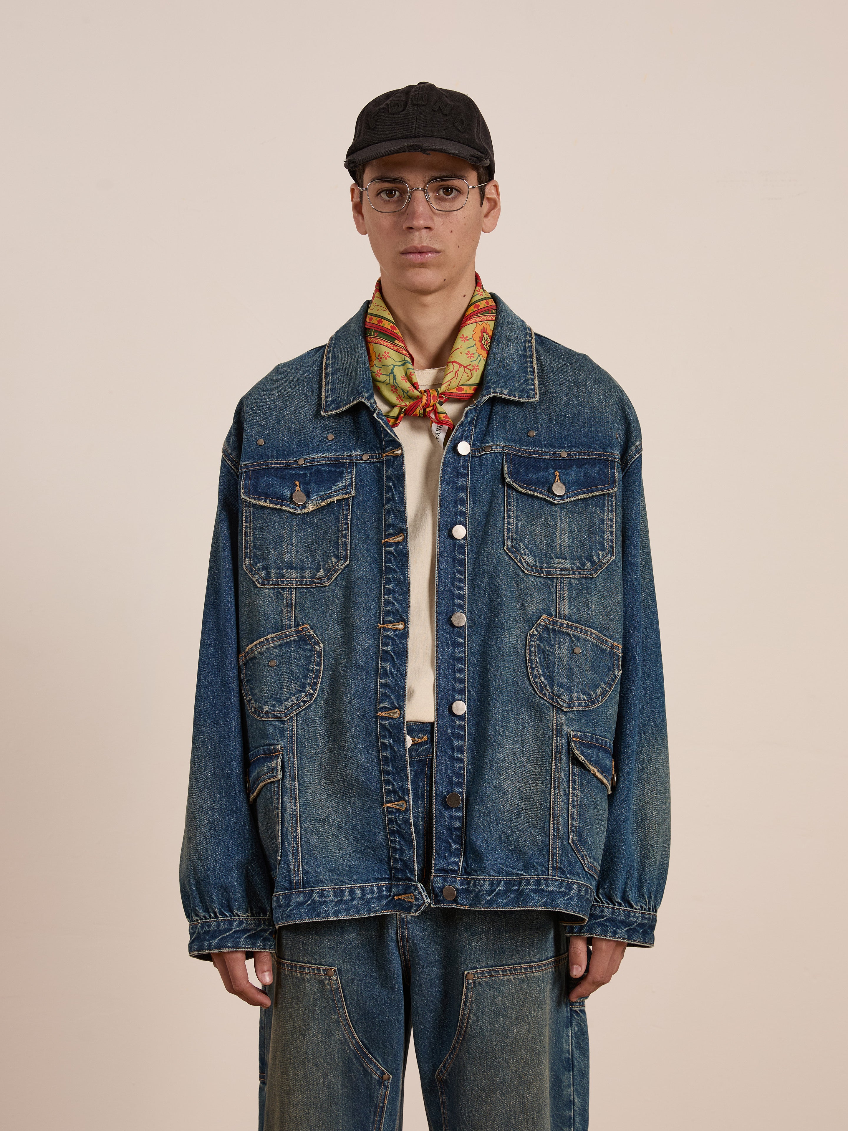 Against a plain background, a person models an effortlessly stylish workwear look in FOUND's unisex relaxed fit Denim Carpenter Jacket and jeans. A colorful scarf adds flair, while the cap and glasses complete the trendy ensemble.