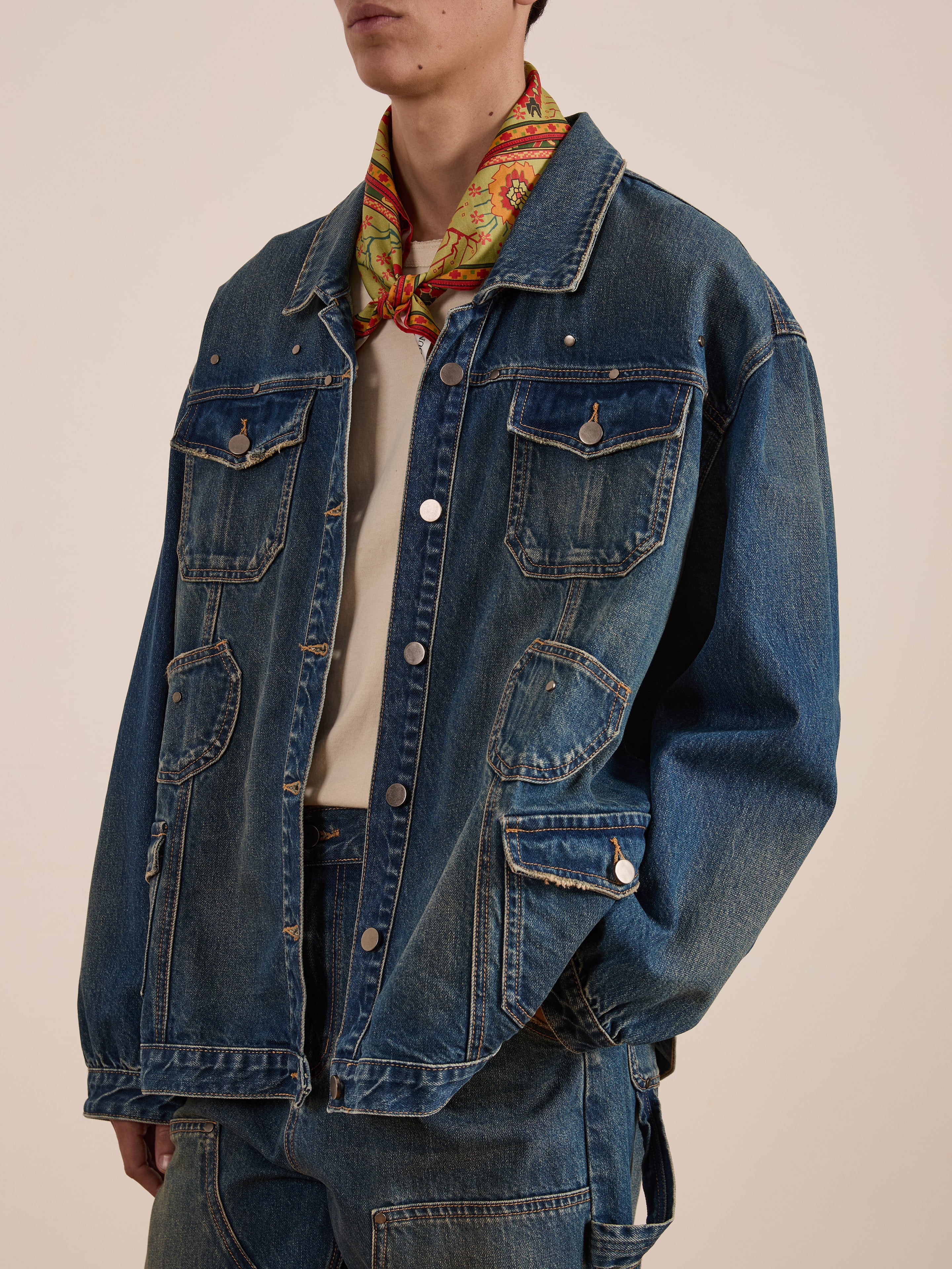 A person wearing a FOUND blue Denim Carpenter Jacket, jeans, a vibrant scarf, and a white shirt in a unisex relaxed fit stands against a plain background.