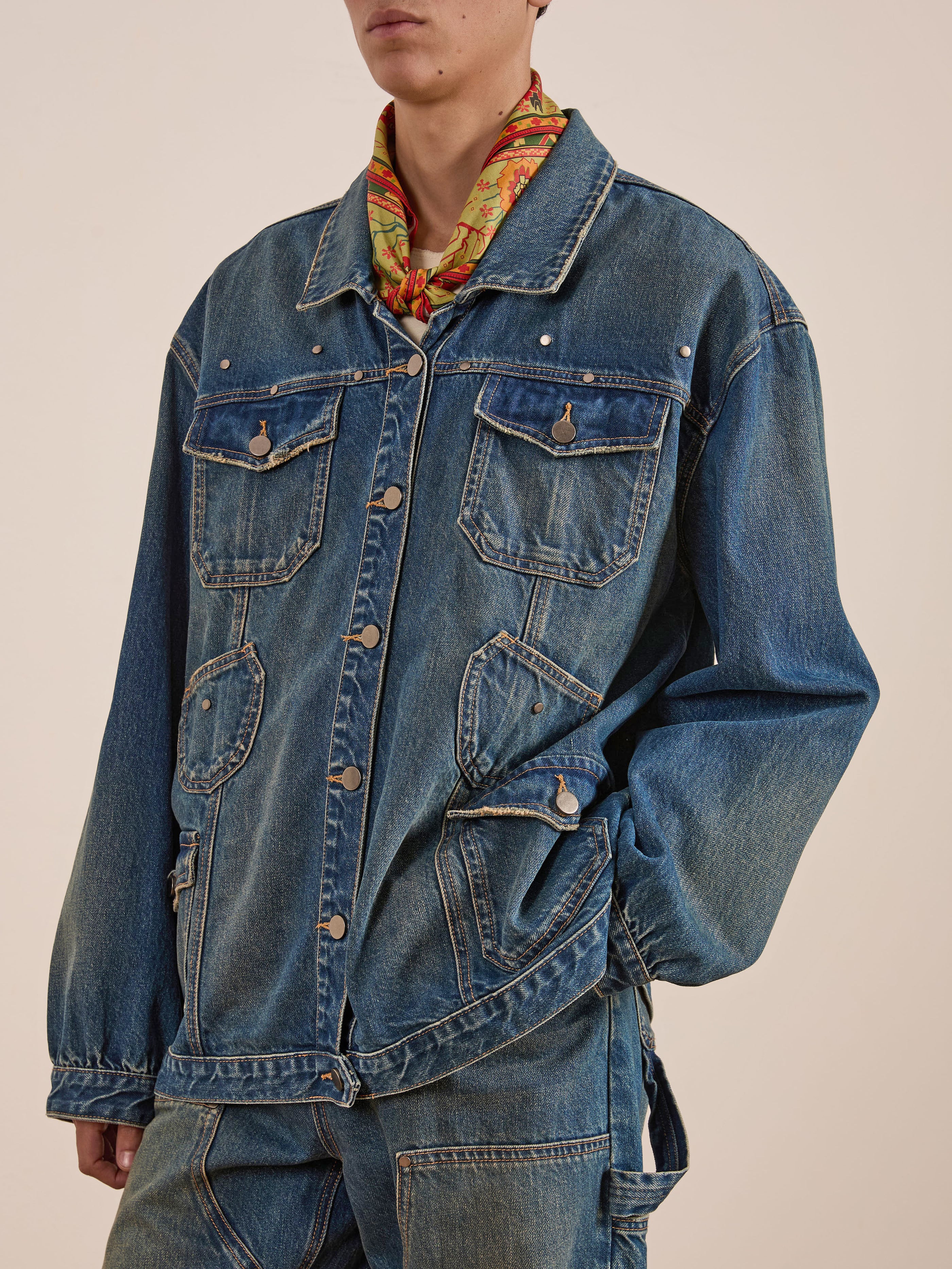 A person dons a FOUND Denim Carpenter Jacket, a workwear staple with utility, buttons, and chest pockets. It stylishly layers over a vibrant scarf.