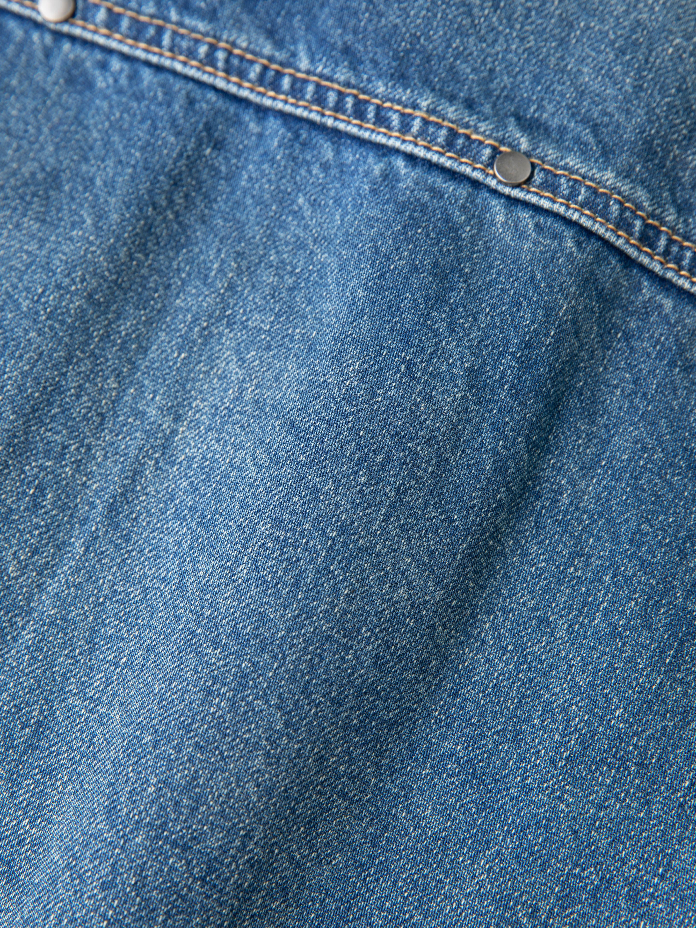 A close-up of blue denim fabric with a seam highlighted by two metal rivets captures the rugged texture of a workwear staple, embodying the spirit of FOUND's Unisex Relaxed Fit Denim Carpenter Jacket.