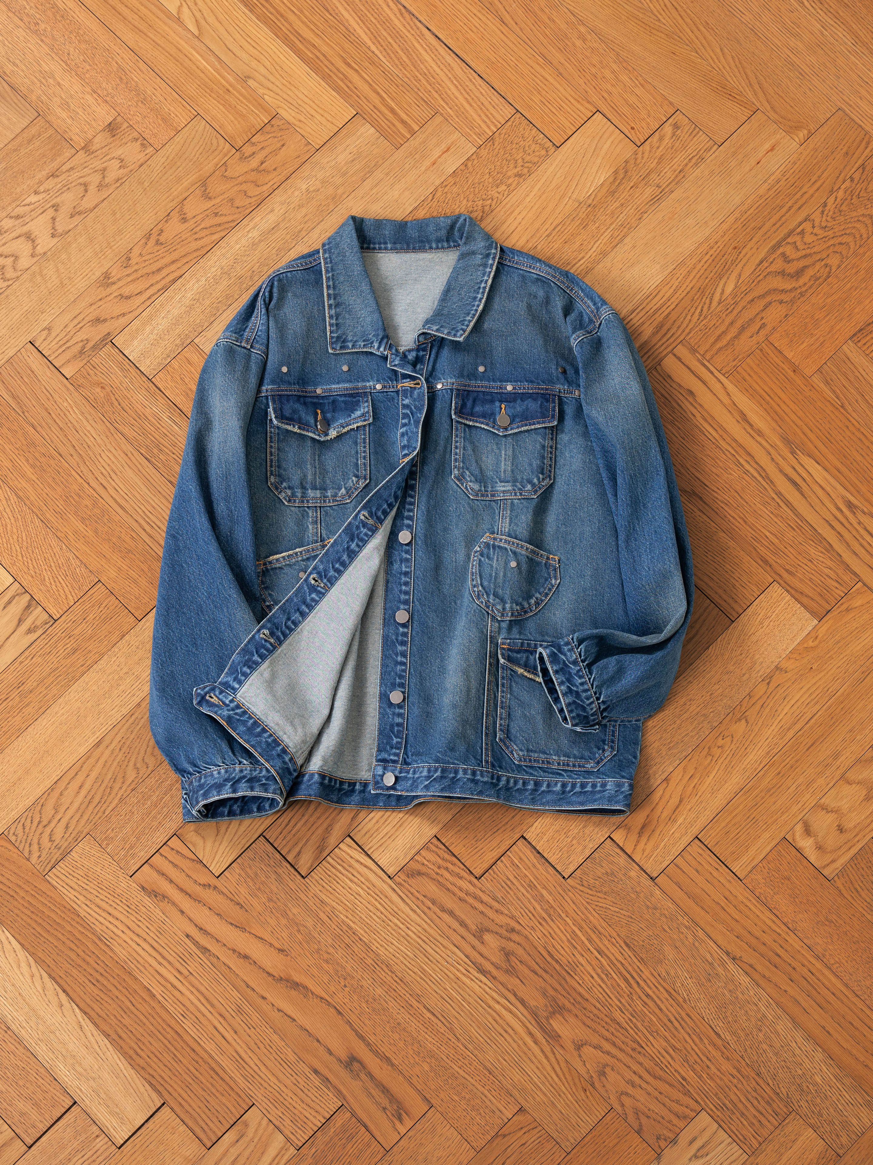 The FOUND Denim Carpenter Jacket, a unisex relaxed fit with buttons and four pockets, is laid on a wooden herringbone-pattern floor, offering the perfect versatile workwear staple.