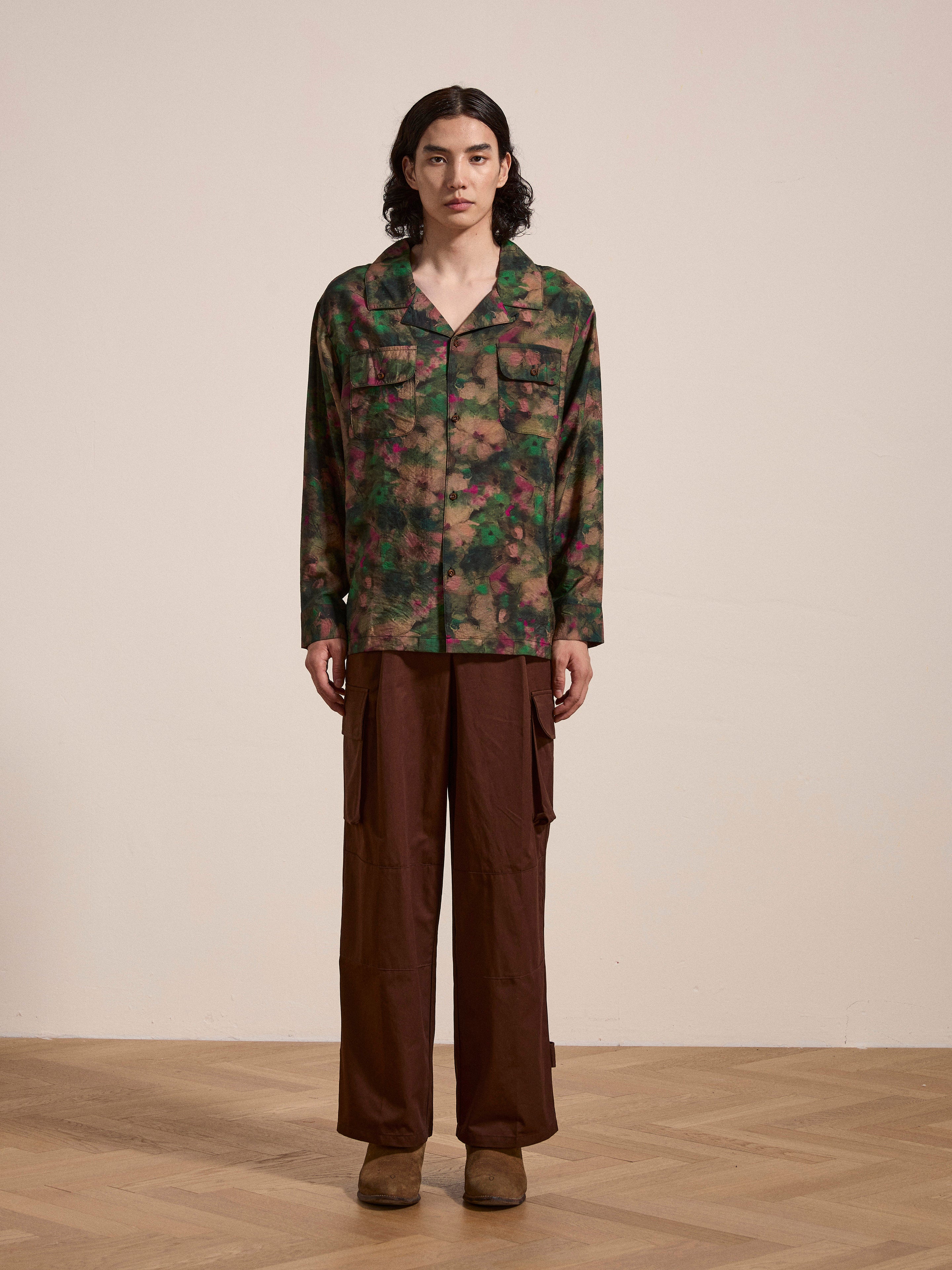A person with long hair stands on a wooden floor, wearing a Charlot LS Camp Shirt by FOUND, featuring dual chest pockets in a green and brown camouflage pattern, paired with brown wide-leg pants and brown shoes. They are looking directly at the camera.