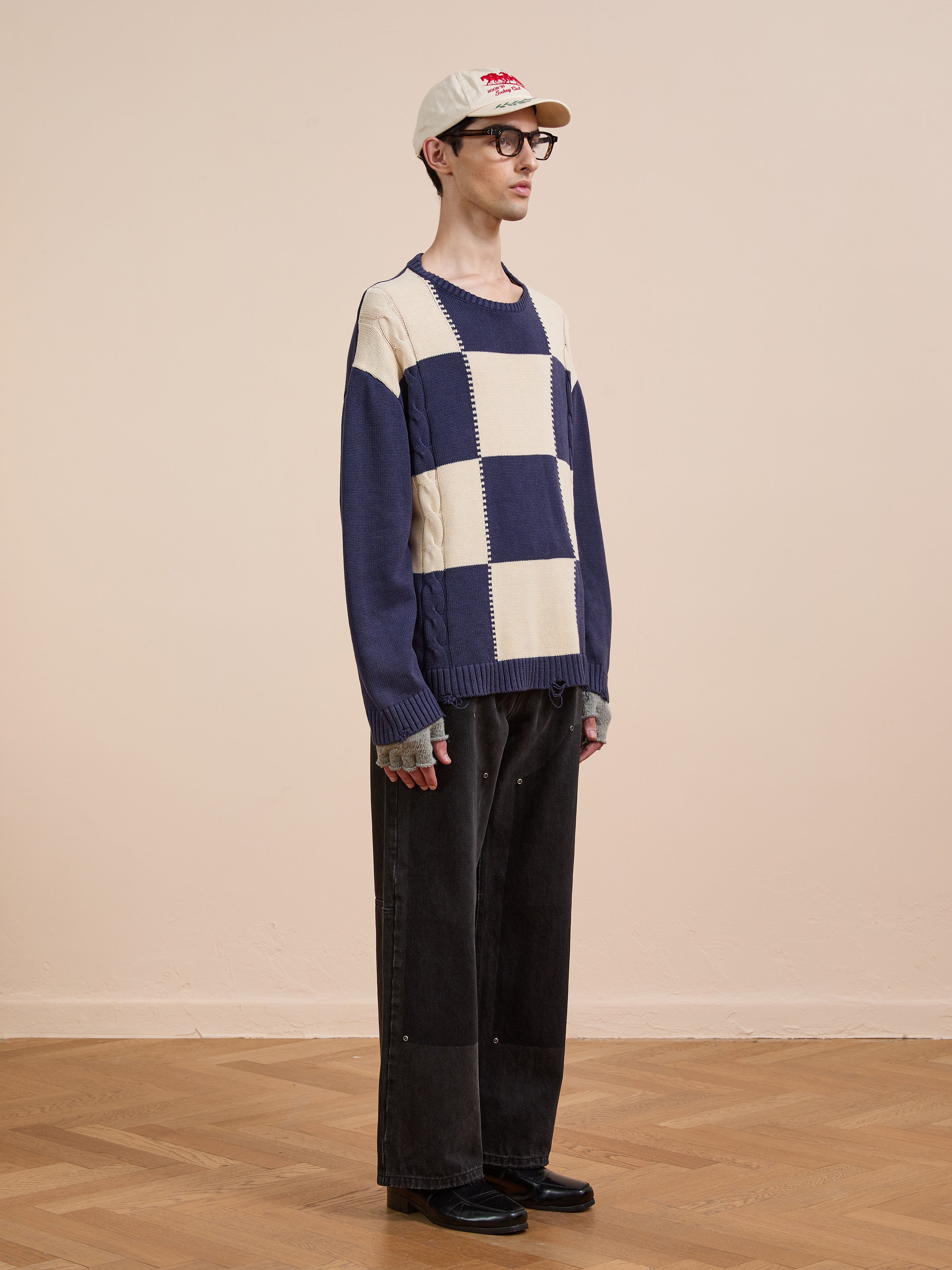 A person stands indoors against a beige wall wearing Found's relaxed fit, cream and navy Checkered Fisherman Sweater, paired with wide-leg black pants, black shoes, glasses, and a white cap.