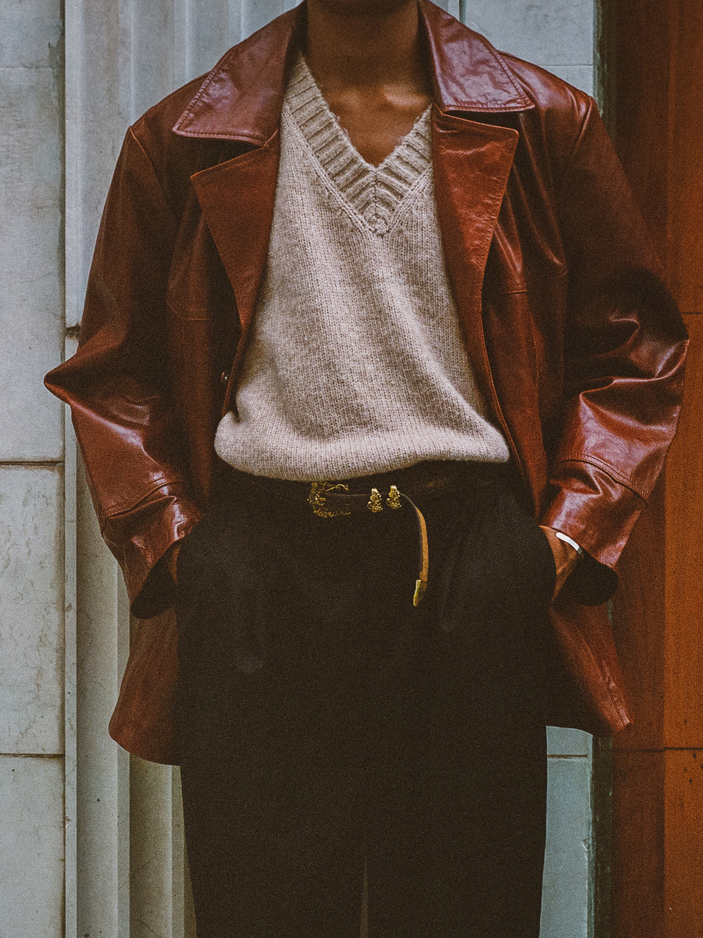 Dressed in a FOUND Claret Western Oversized Leather Blazer over a beige sweater and black pants, the person confidently stands in front of a building facade.