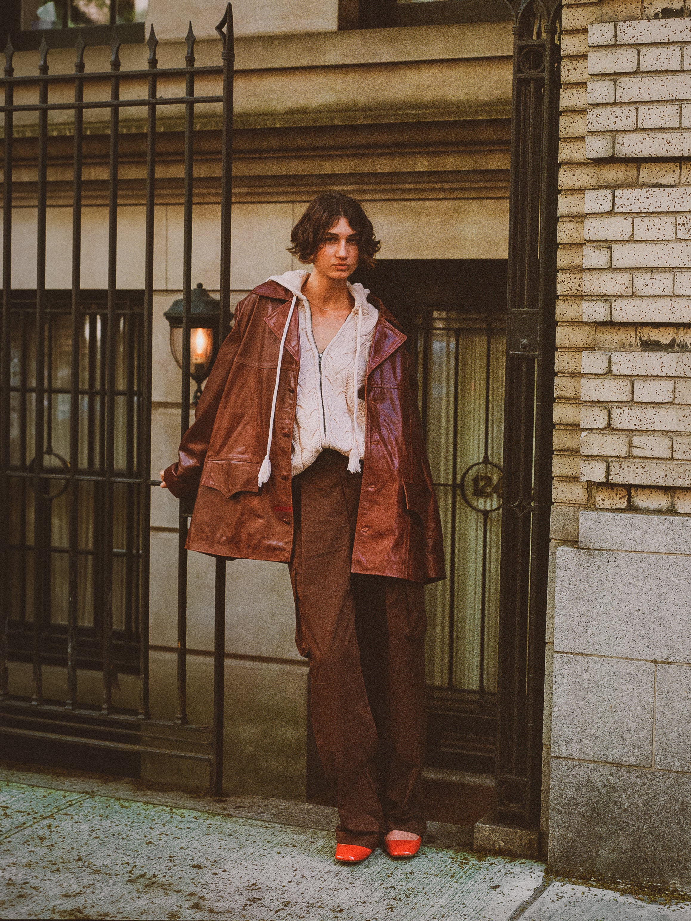 In an urban setting, a person with short hair stands by an iron fence and brick wall, wearing the FOUND Claret Western Oversized Leather Blazer over a white shirt, paired with brown pants and red shoes.