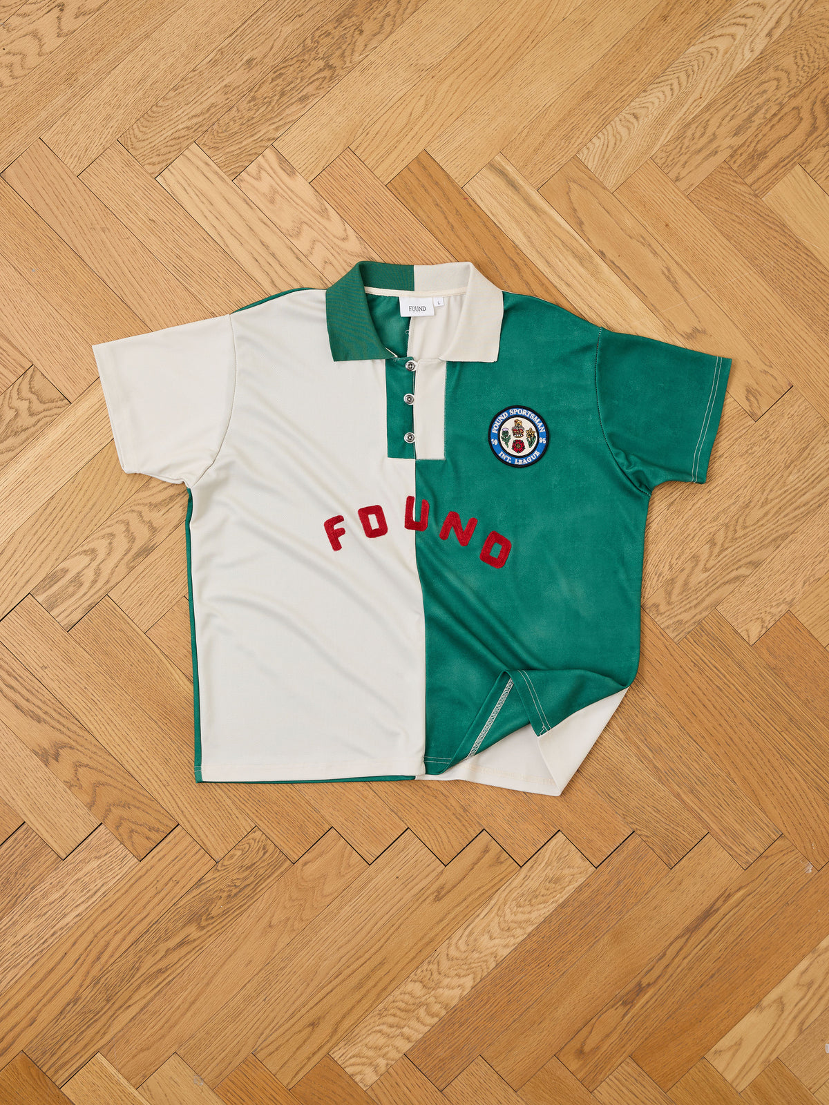 The FOUND Color Block Regent Jersey is a short-sleeve, collared collegiate polo featuring bold vertical color blocking in white and green halves, accented by “FOUND” in red lettering and a round embroidered chest patch—a true nod to vintage sportswear style, shown here lying flat on a wooden floor.