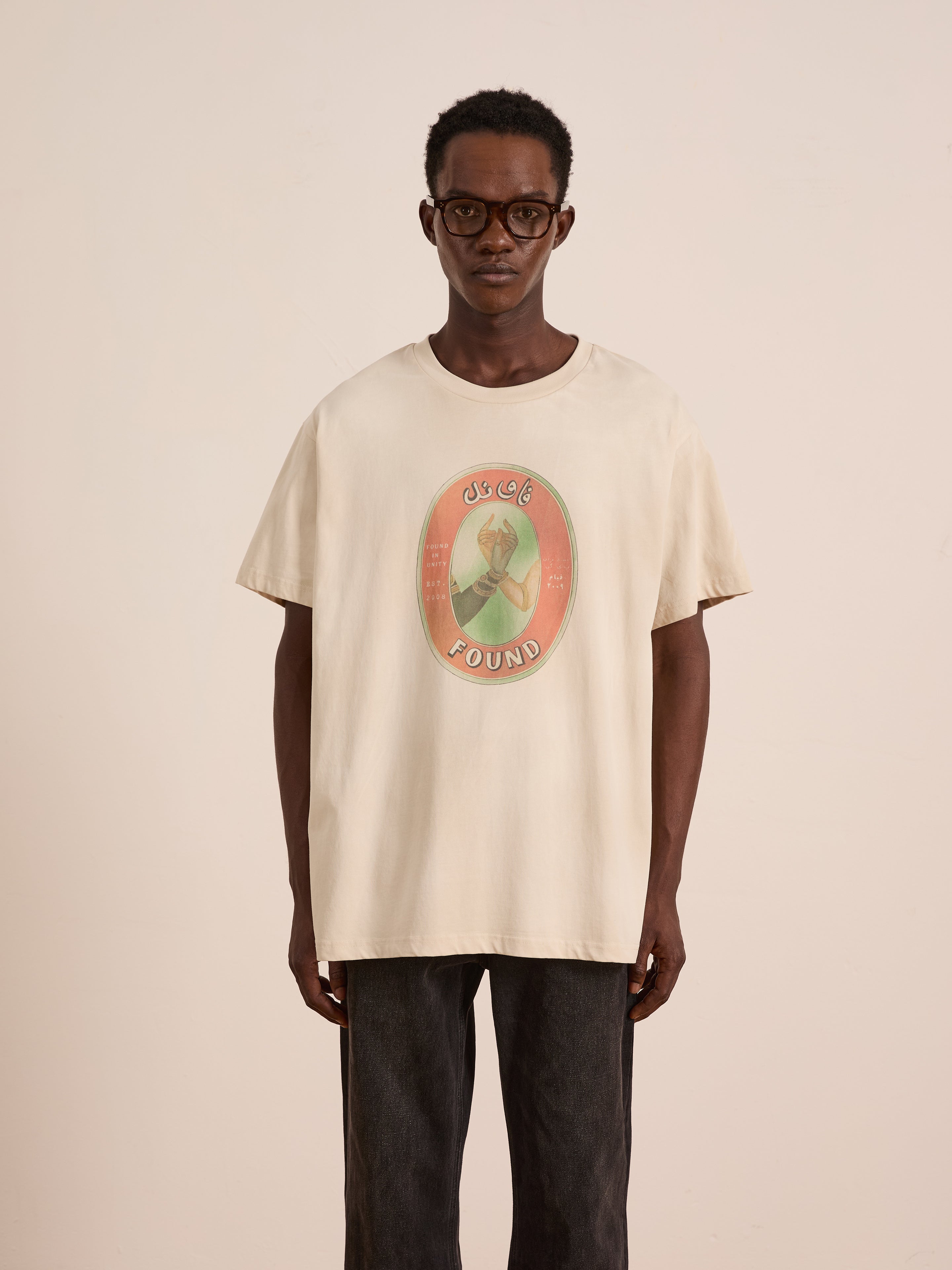 A man wearing glasses and dark pants stands against a light background in FOUND’s Community Hands Tee—a beige unisex cotton shirt with a circular graphic and "LOST & FOUND," inspired by vintage matchbox art.
