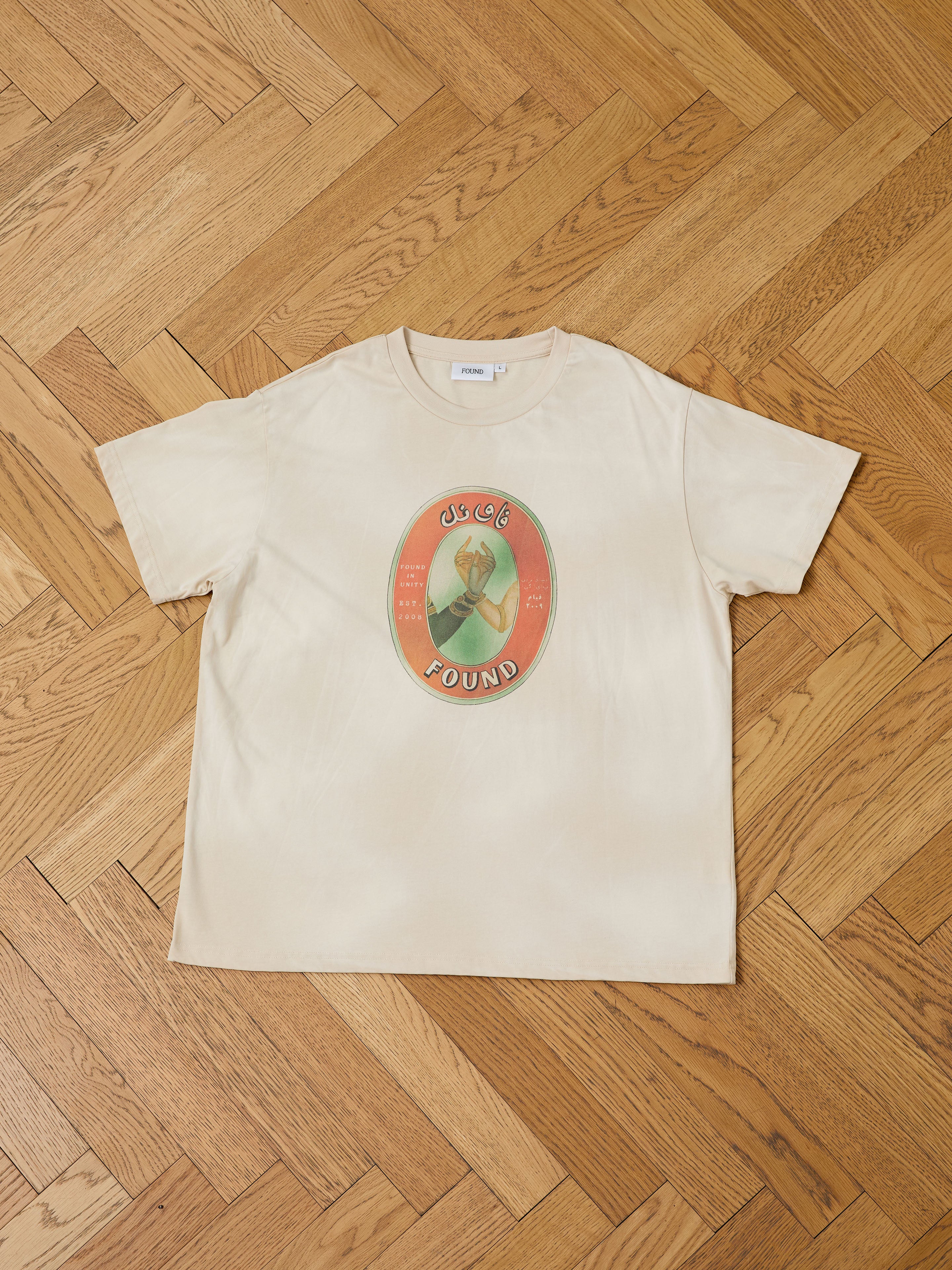 The Community Hands Tee by FOUND is a beige unisex cotton shirt with a vibrant matchbox-inspired oval graphic, showing a raised fist, Arabic script, and FOUND branding. Pictured laid flat on a herringbone wood floor.