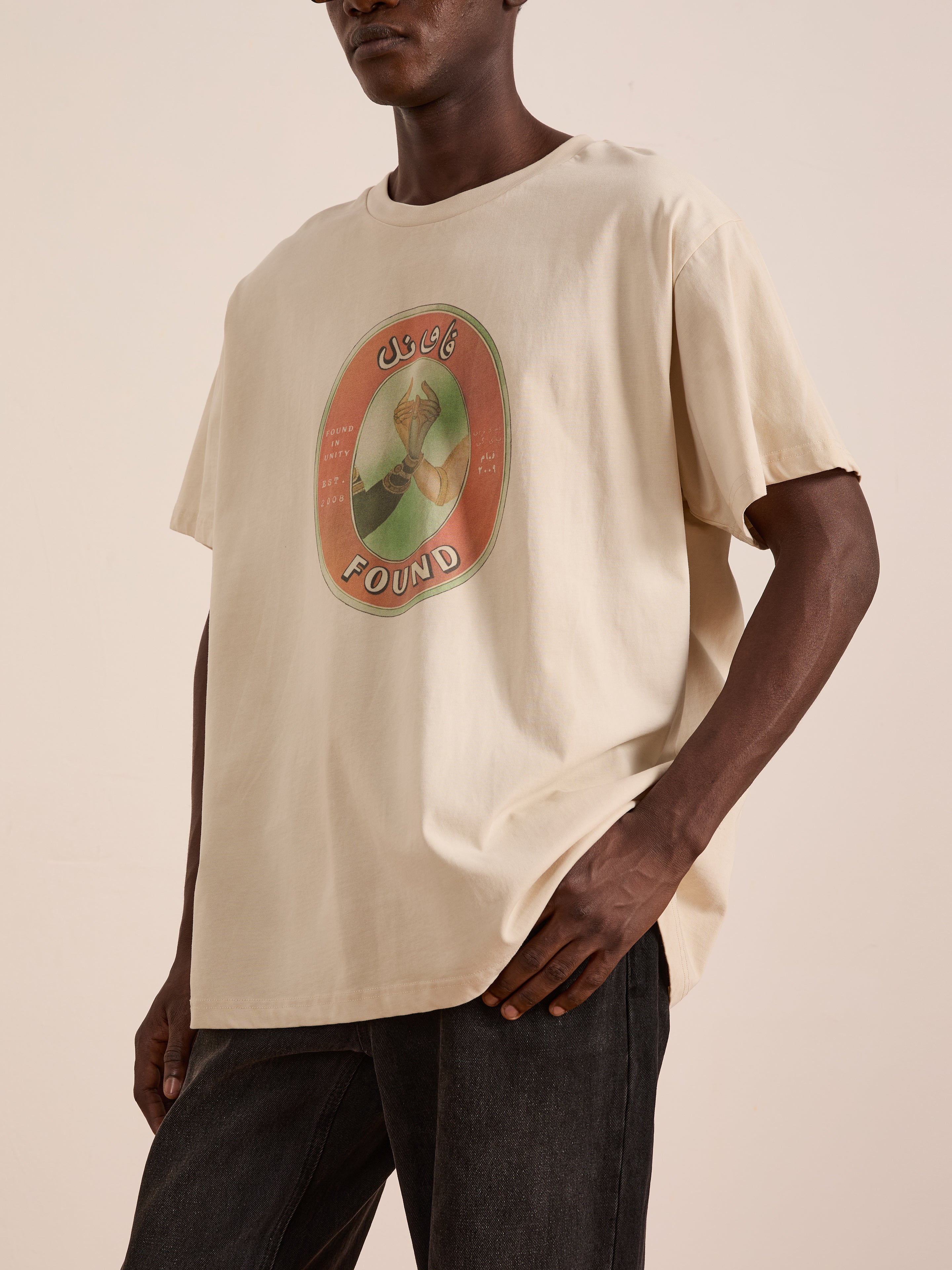 Someone wears FOUND’s Community Hands Tee, a loose unisex beige cotton t-shirt with a multicolored oval graphic and "FOUND IN UNITY" on the front, inspired by vintage matchbox art, paired with dark pants.