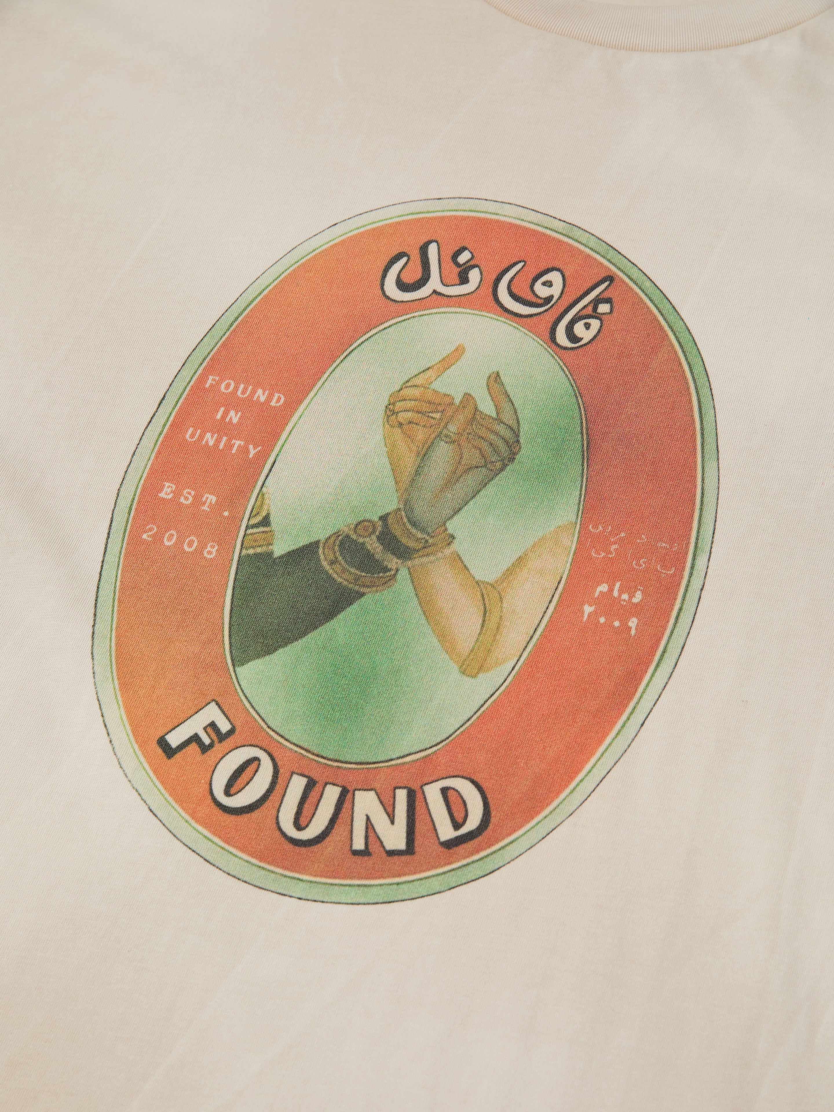 The FOUND Community Hands Tee features two hands reaching inside an oval, blending vintage matchbox art with English and Arabic text: "Found in Unity Est. 2008" and "FOUND." A unique t-shirt statement about unity.