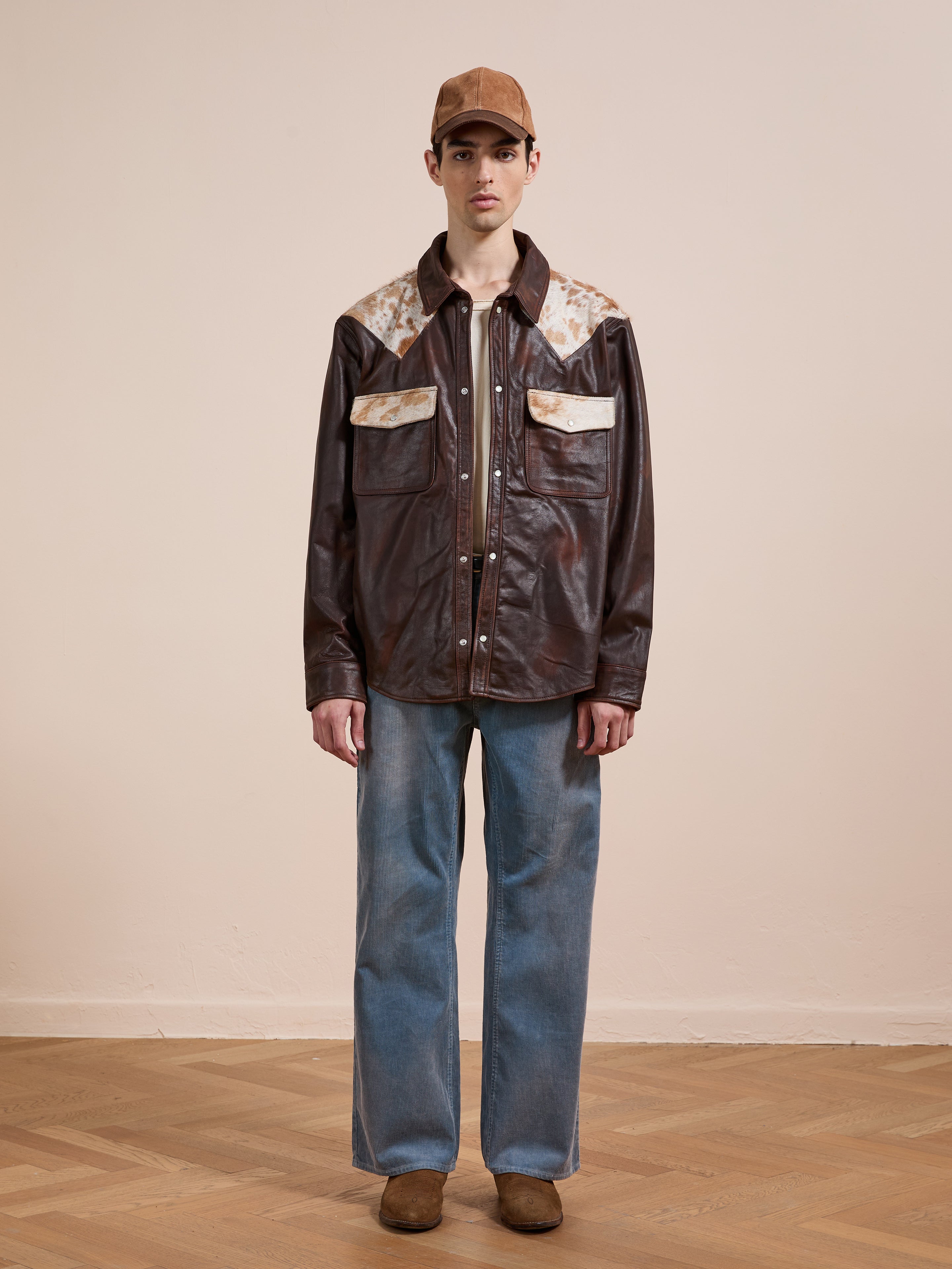 A man stands indoors facing forward against a plain light background, wearing the FOUND Distressed Leather Cowhide Contrast Western Shirt, blue jeans, brown shoes, and a brown cap.