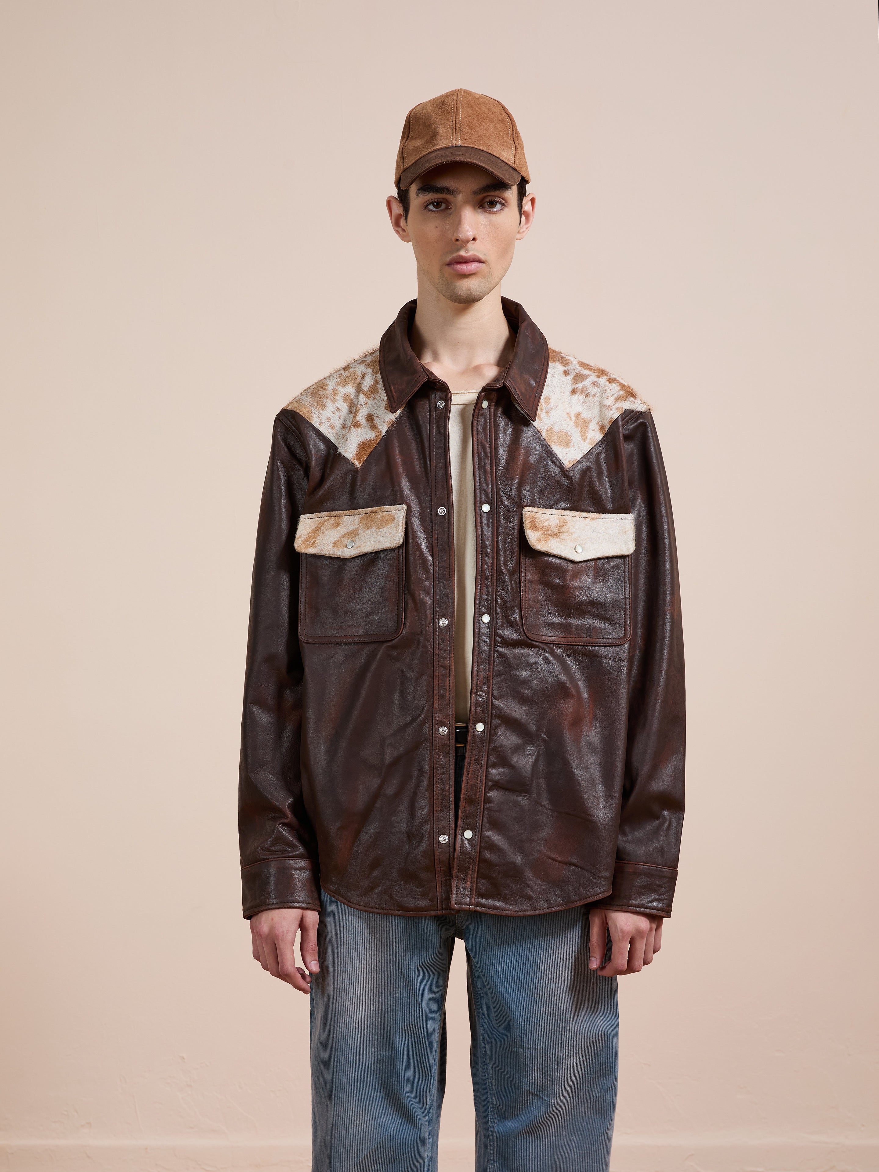 A person wearing a FOUND Distressed Leather Cowhide Contrast Western Shirt, blue jeans, and a brown cap stands against a plain light-colored background.