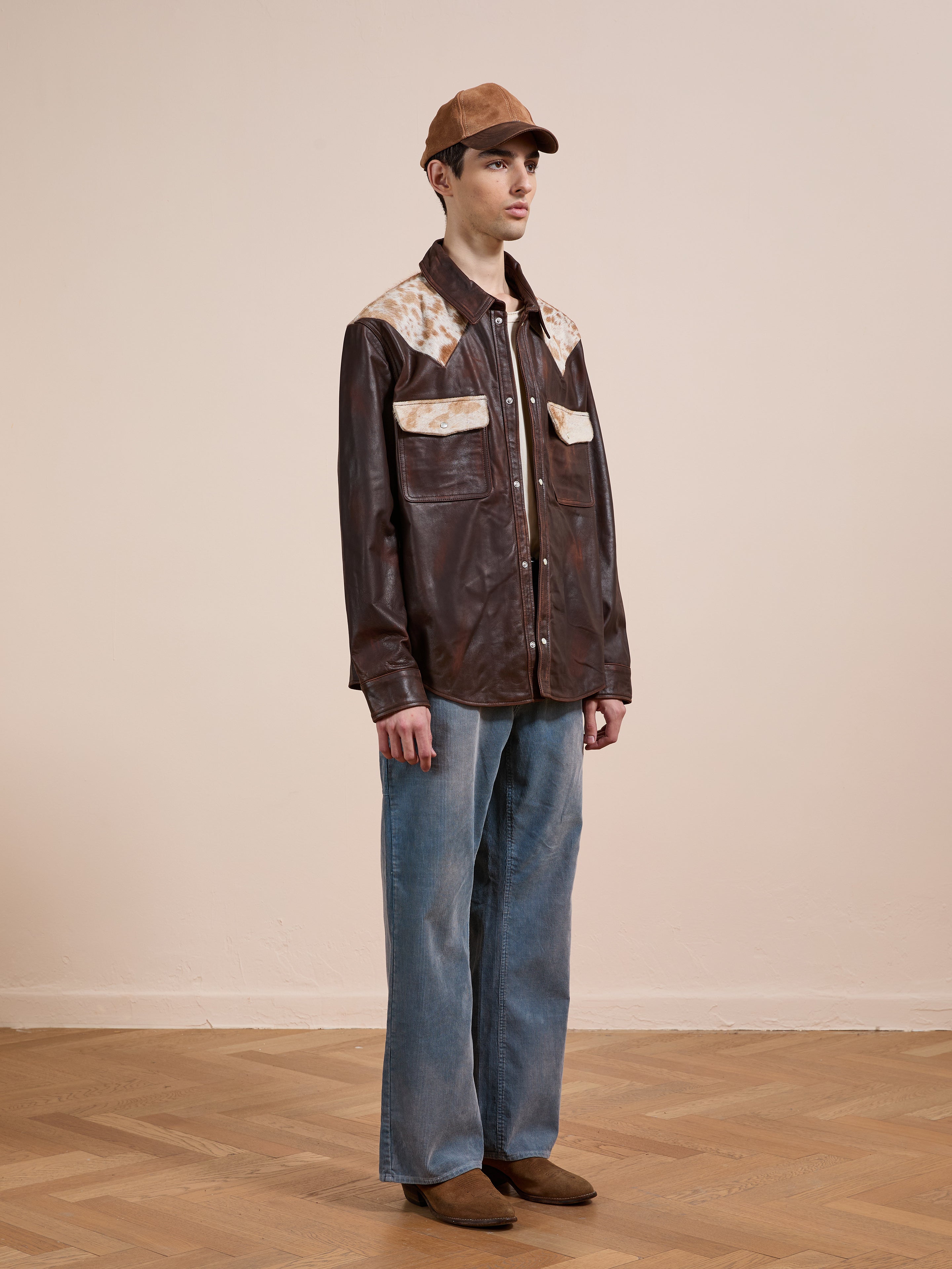 A person stands indoors against a plain light wall wearing the FOUND Distressed Leather Cowhide Contrast Western Shirt, blue jeans, brown shoes, and a brown cap.