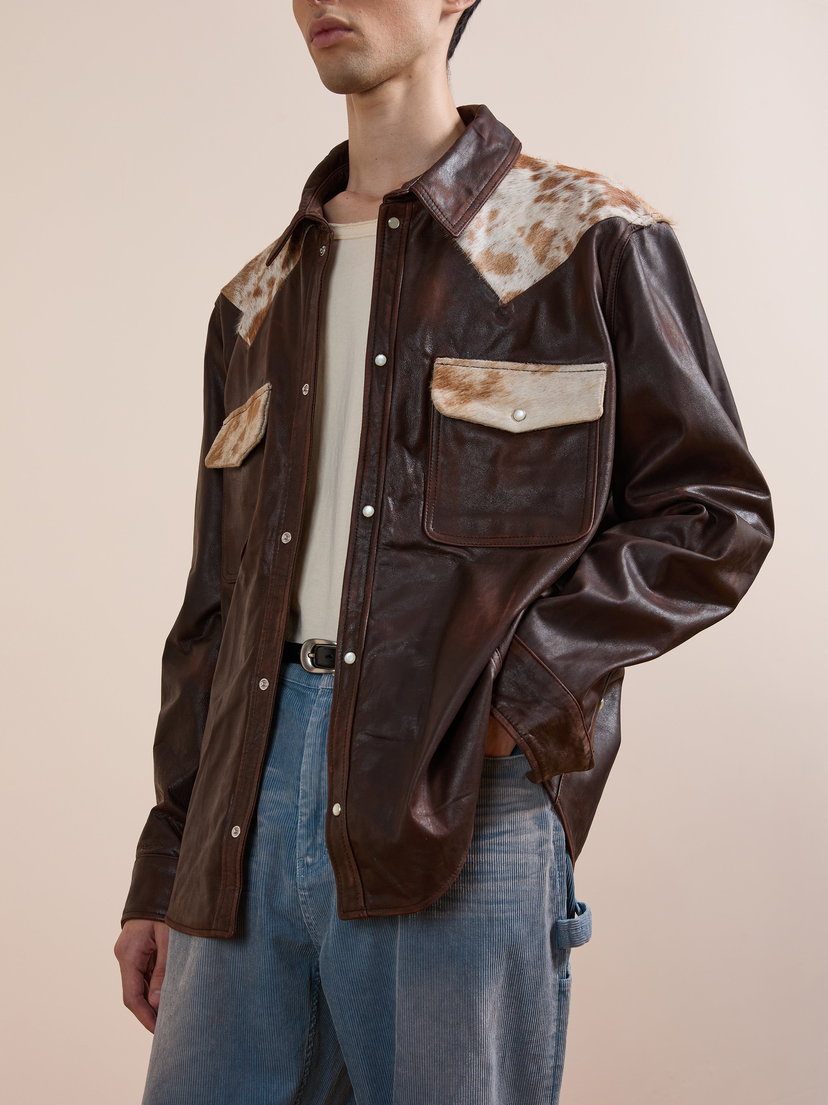 A person wears the FOUND Distressed Leather Cowhide Contrast Western Shirt with fur-accented shoulders and pockets, a beige shirt, and light blue jeans against a plain background.