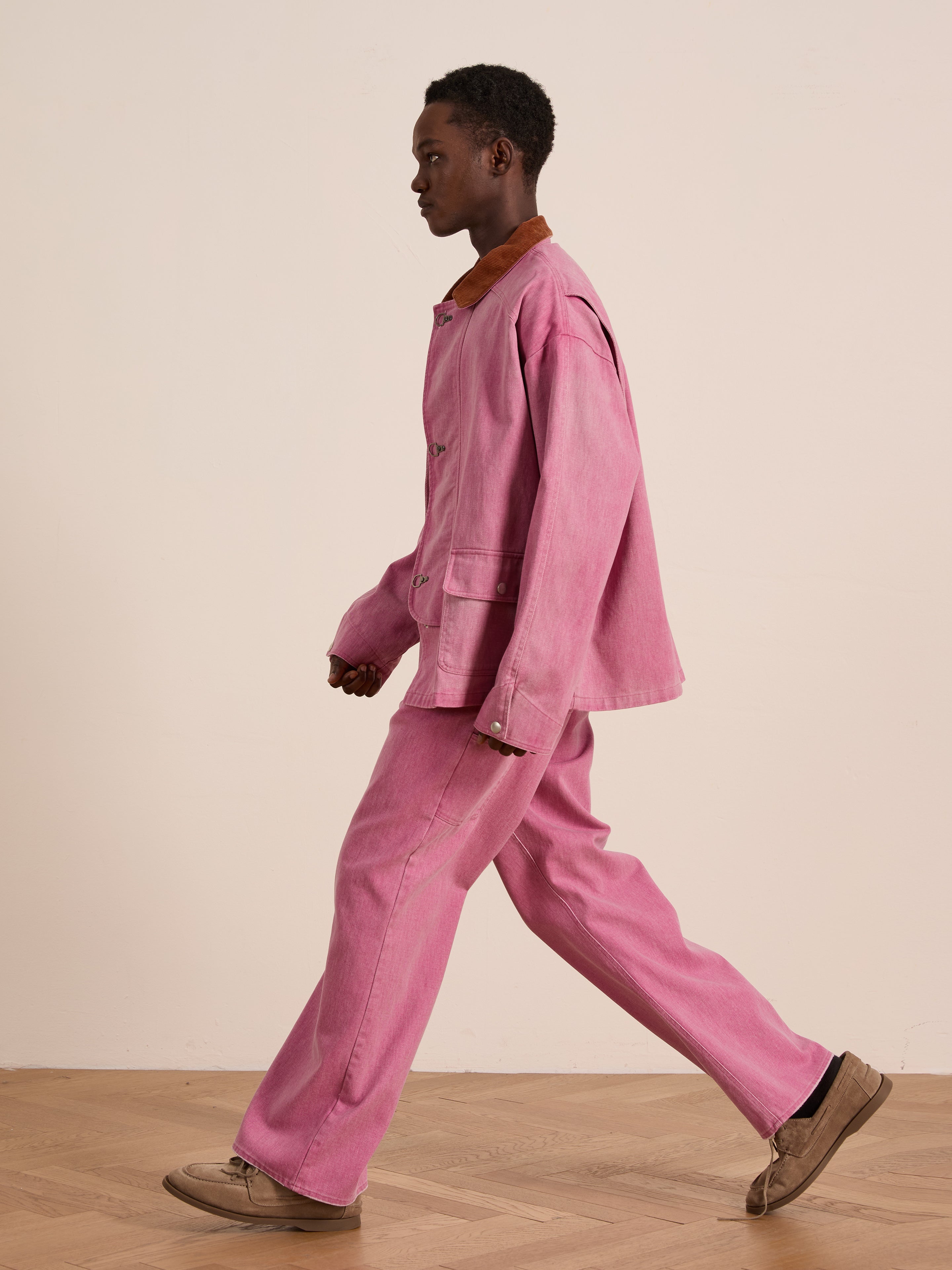 A person walks indoors wearing Found’s Washed Fireman Clasp Workshop Jacket in pink with matching pants and tan shoes, set against a plain light-colored wall on a wooden floor.