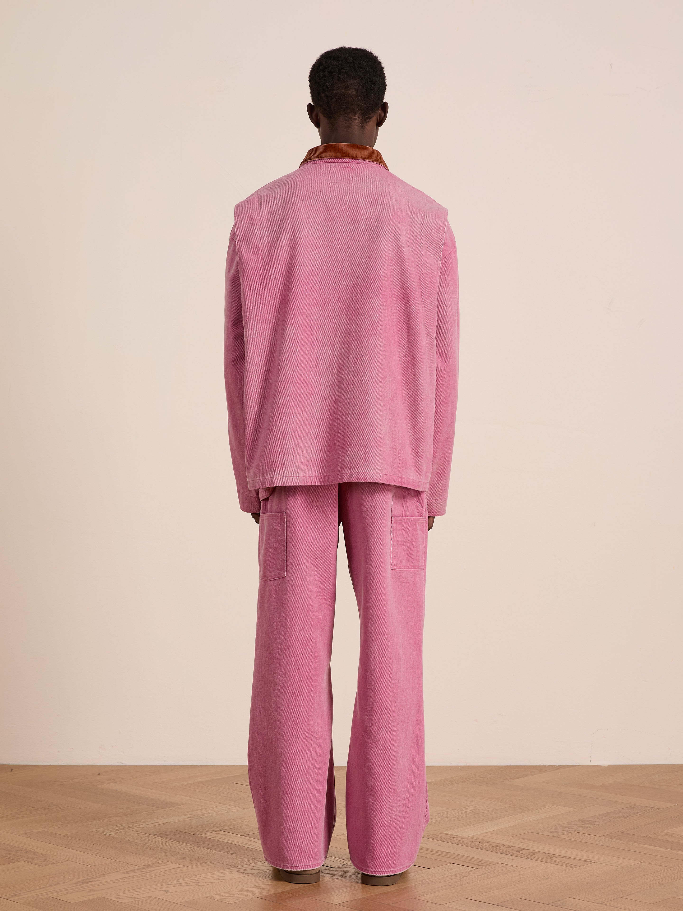 A person faces away from the camera, wearing the Found Washed Fireman Clasp Workshop Jacket in pink, paired with matching pants, on a wooden floor against a plain beige wall.