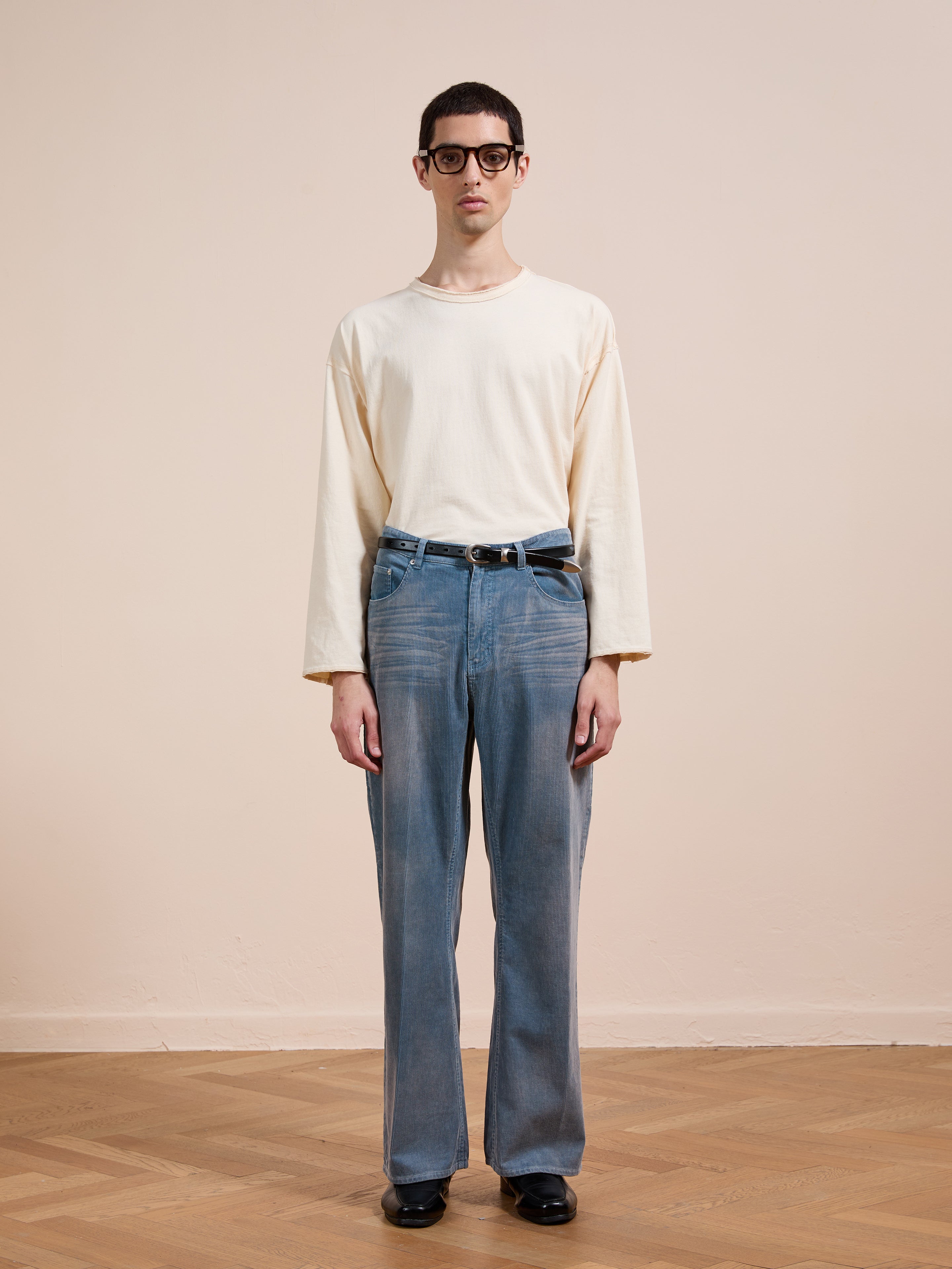A person wears black glasses, an off-white long-sleeve shirt, FOUND Corduroy Flare Work Pants in vintage blue and a relaxed fit, and black shoes while standing on a wooden floor against a plain beige wall.