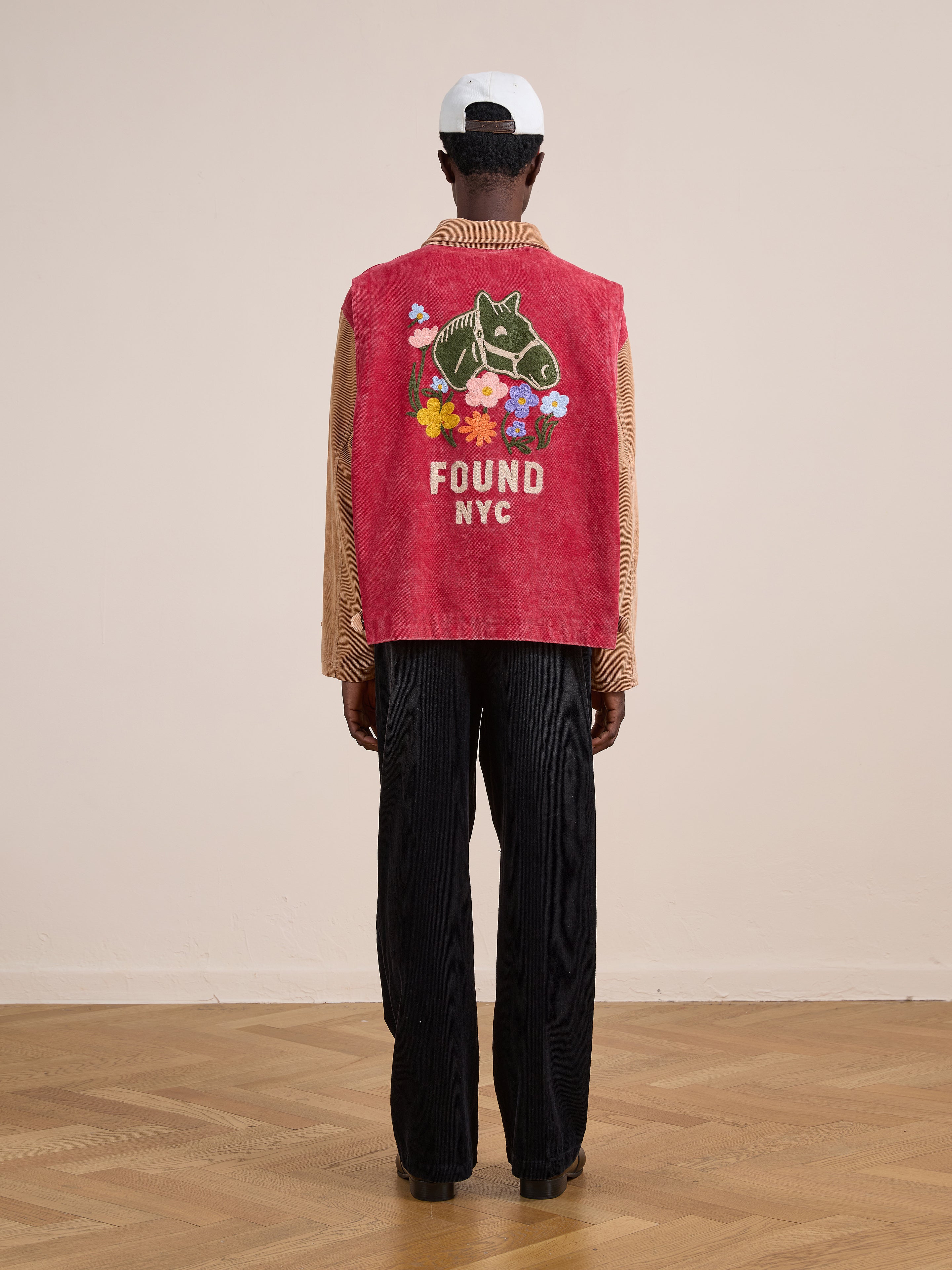 Person standing indoors, facing away, wearing the Found Corduroy Field Jacket in red with a horse graphic, colorful flowers, and "FOUND NYC" text on the back, paired with black pants and a white cap.