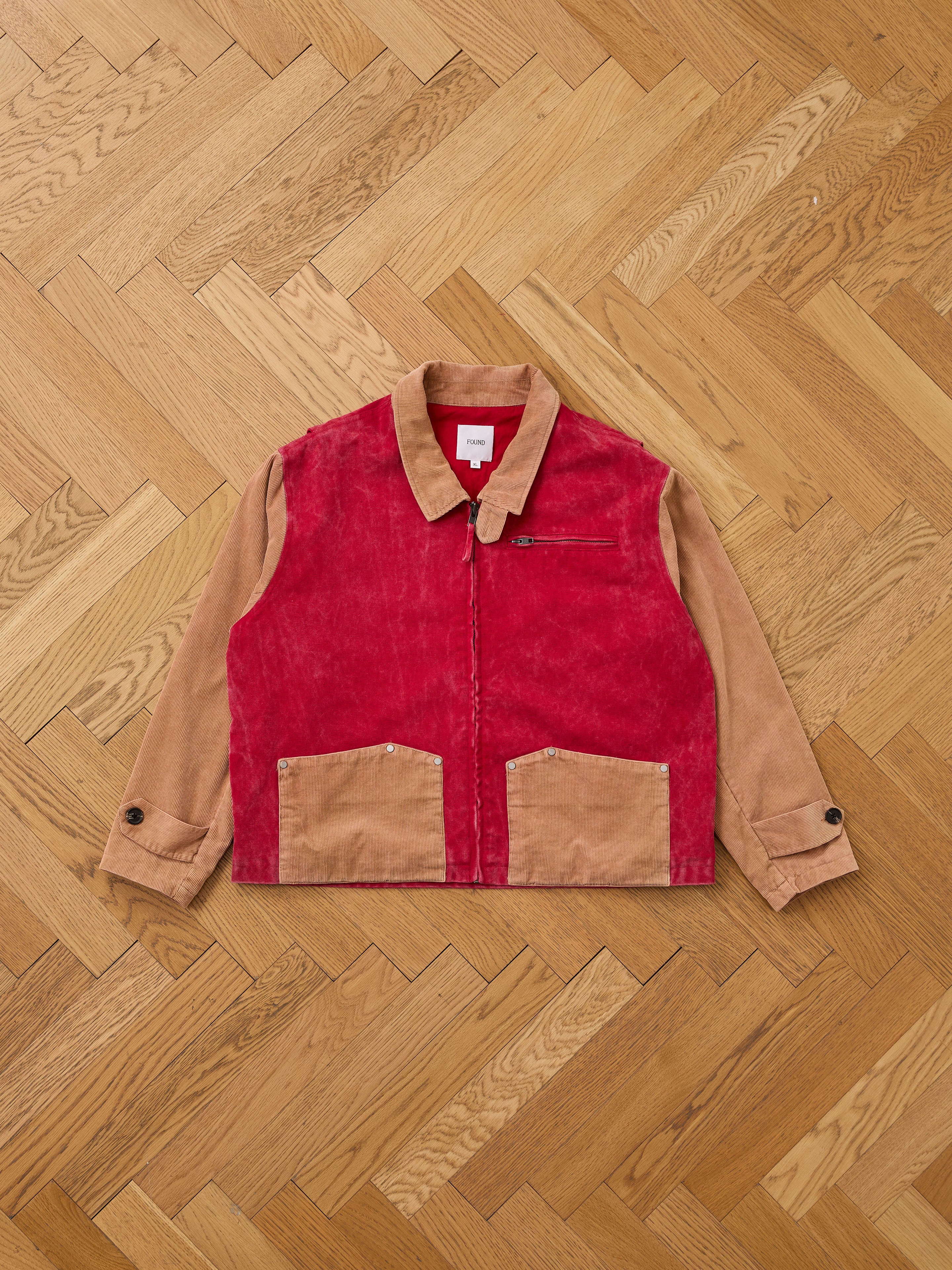 The Found Corduroy Field Jacket features a two-tone design with tan sleeves and collar, a red corduroy front, tan pockets, zippered chest pocket, and buttoned cuffs, shown laid flat on a wooden herringbone floor.