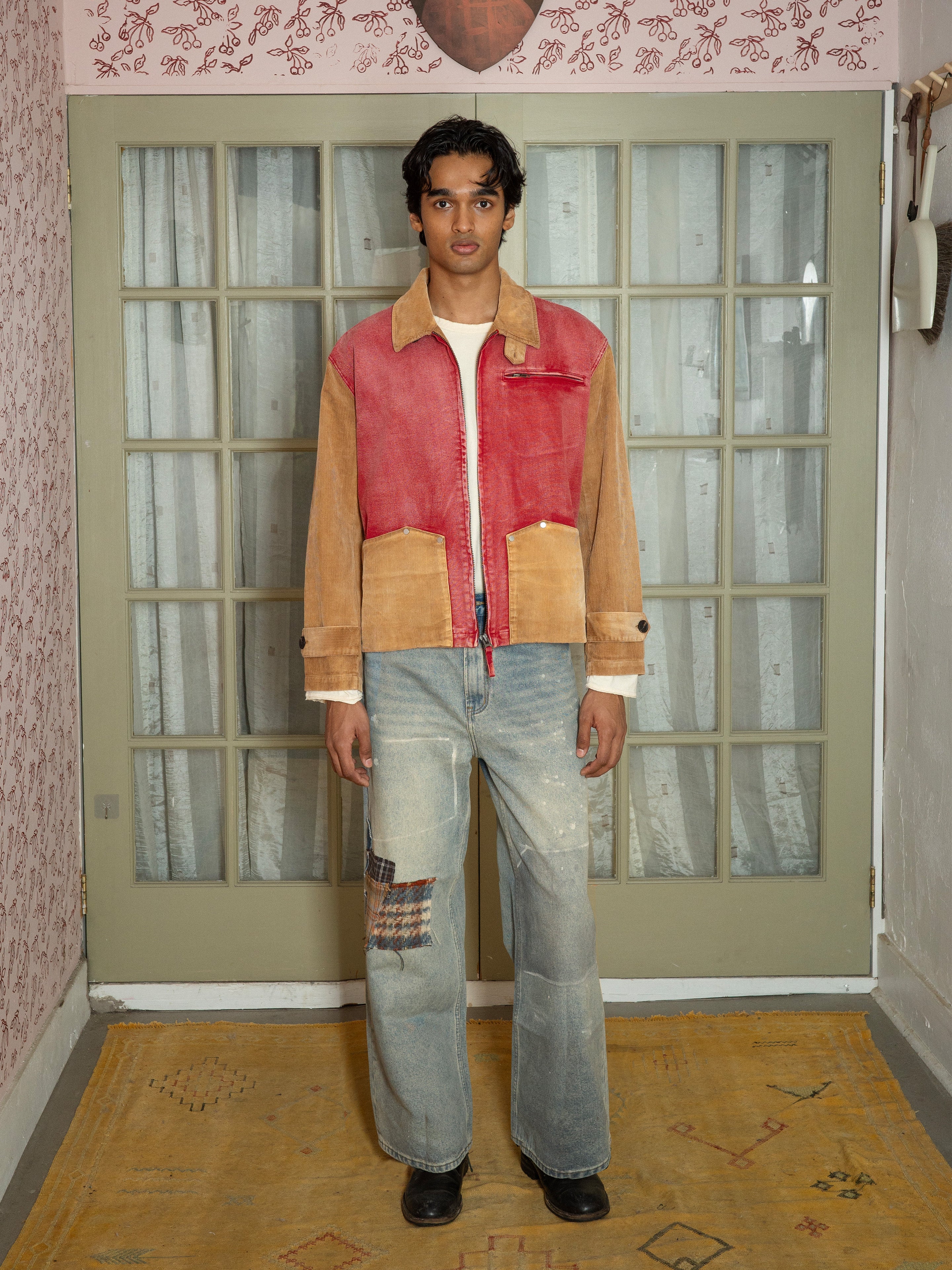 A person stands indoors facing the camera in front of glass doors, dressed in a faded red Found Corduroy Field Jacket, light wash patched jeans, and black shoes. The unisex workwear jacket by Found adds a distinctive touch to the color-blocked ensemble.