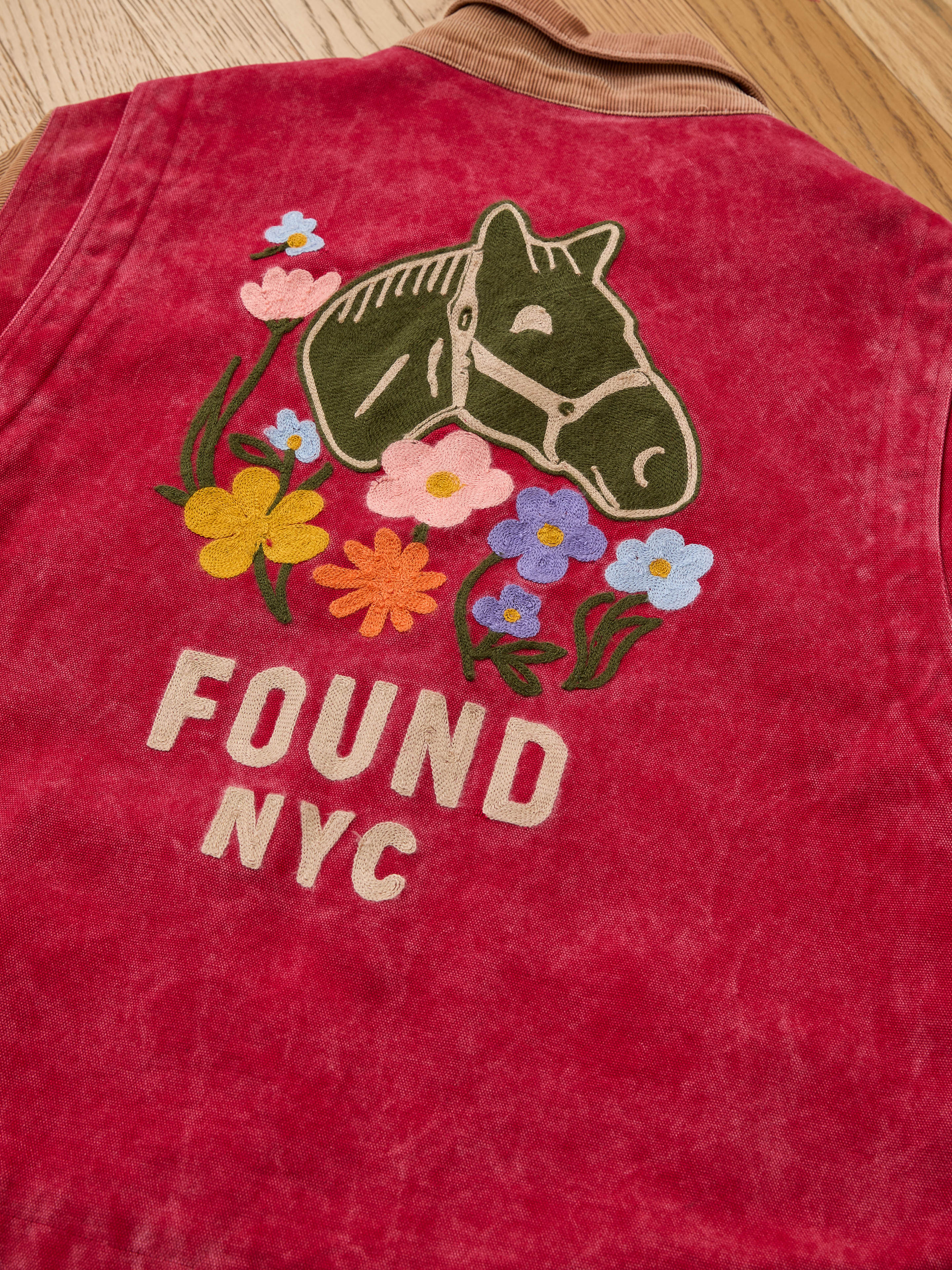 The Found Corduroy Field Jacket is a relaxed-fit unisex piece in red, featuring an embroidered horse head, colorful flowers, and "FOUND NYC" on the back, shown laid flat on wood for a modern take on classic outerwear.