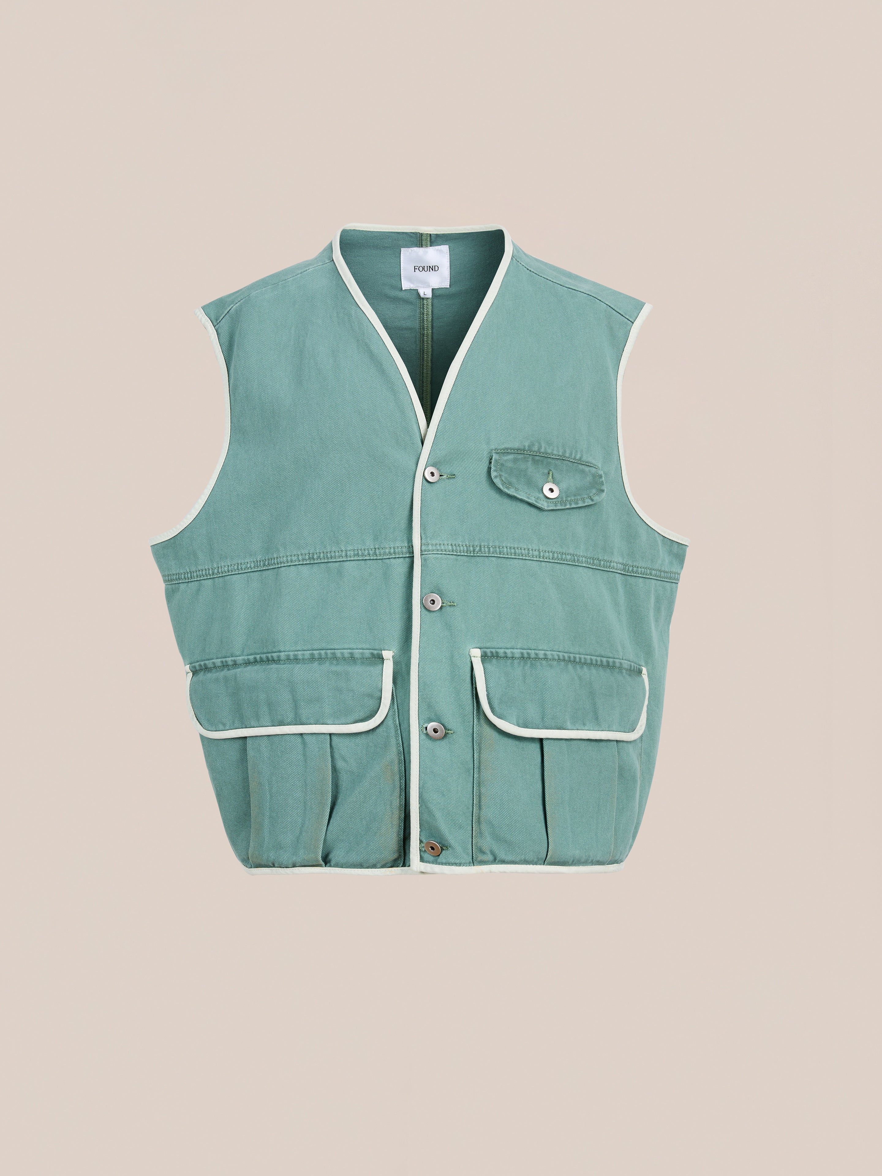 The FOUND Cotton Twill Utility Vest in vintage washed green features a sleeveless design, white piping, front buttons, and three flap pockets, shown against a plain beige background.