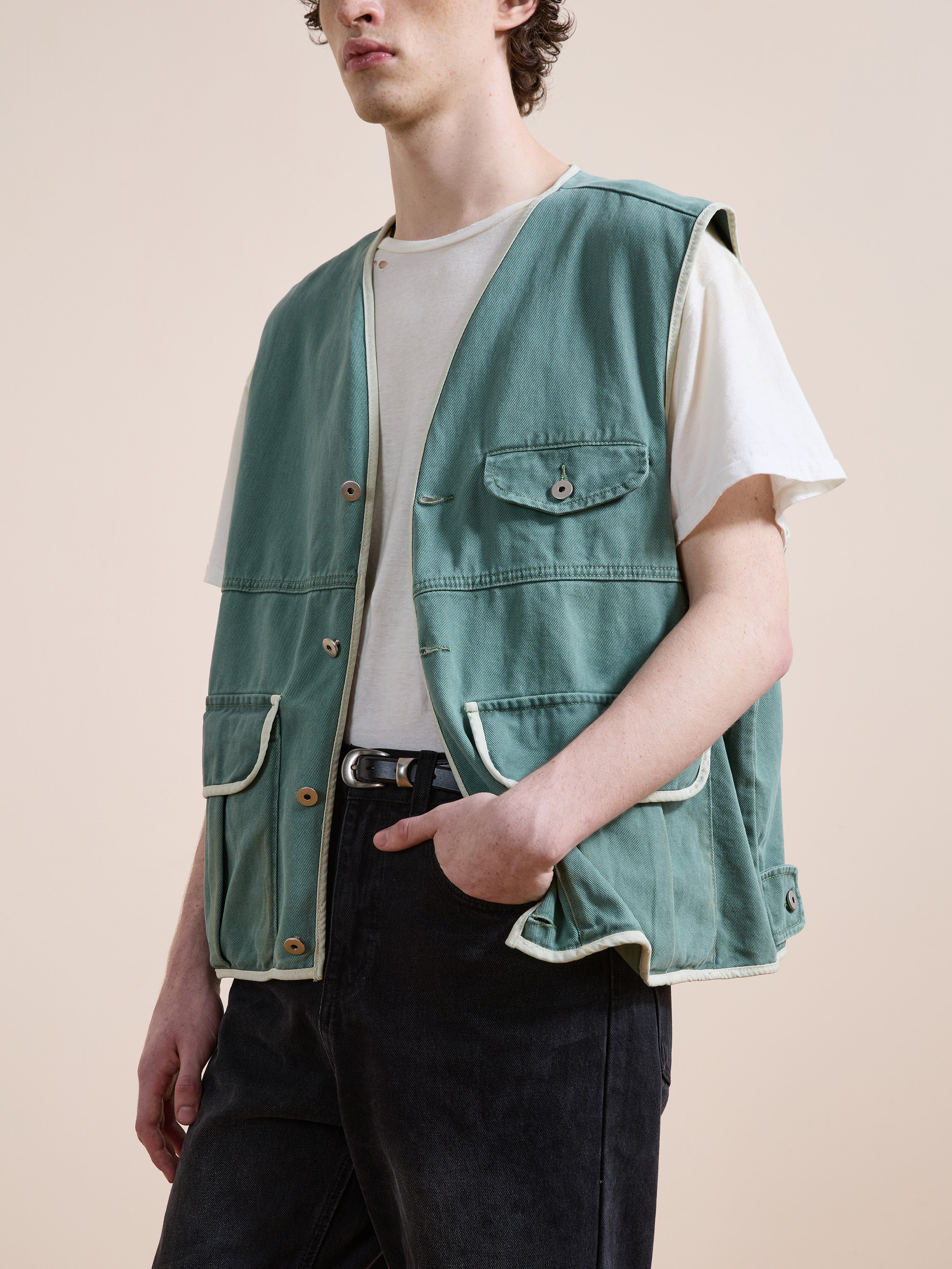A person wearing a white t-shirt, FOUND Cotton Twill Utility Vest in vintage washed green with front pockets, and black jeans stands with one hand in their pocket against a neutral background.