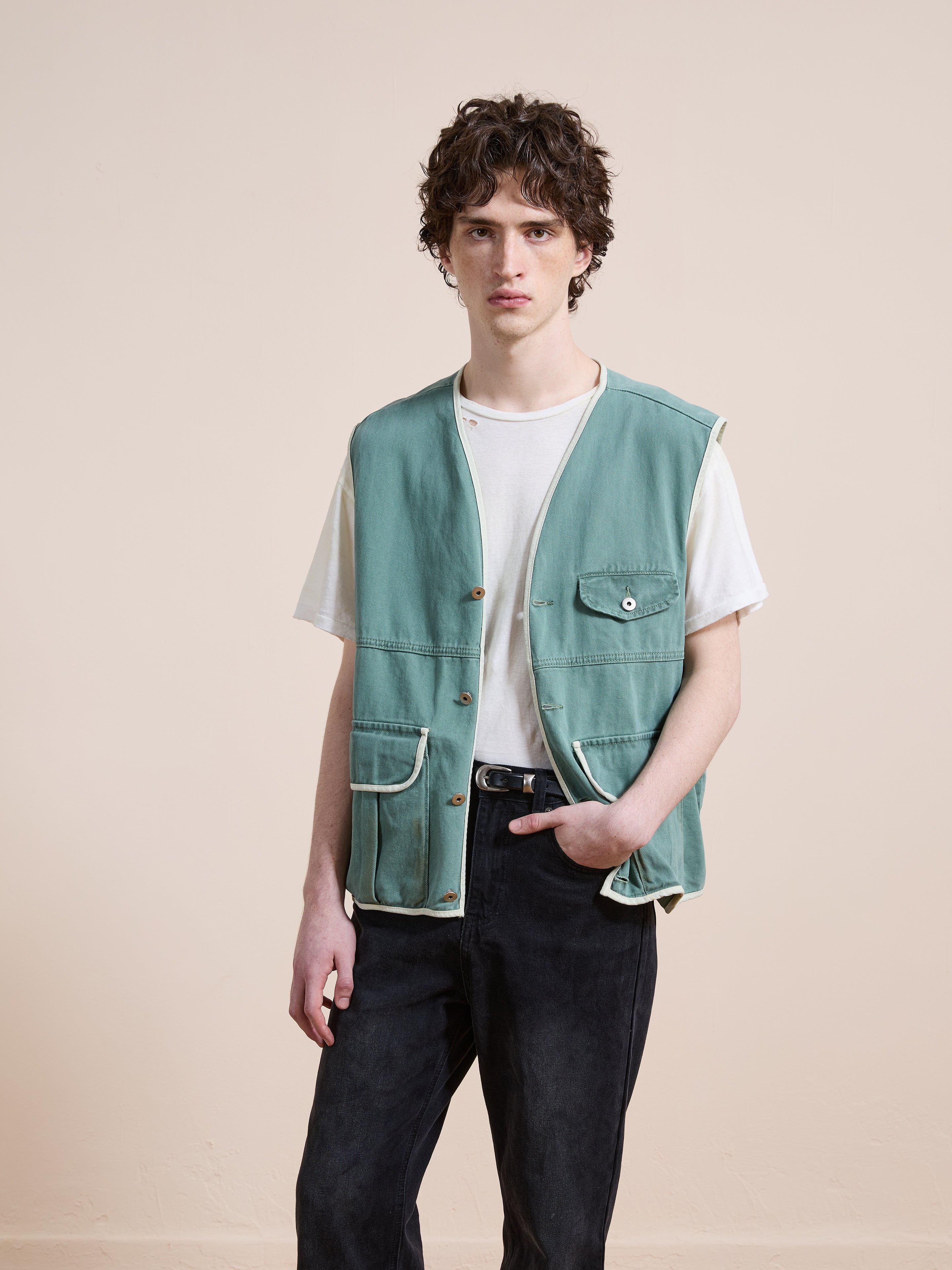 A person wears the FOUND Cotton Twill Utility Vest in vintage green over a white t-shirt and dark pants, standing against a plain beige background.