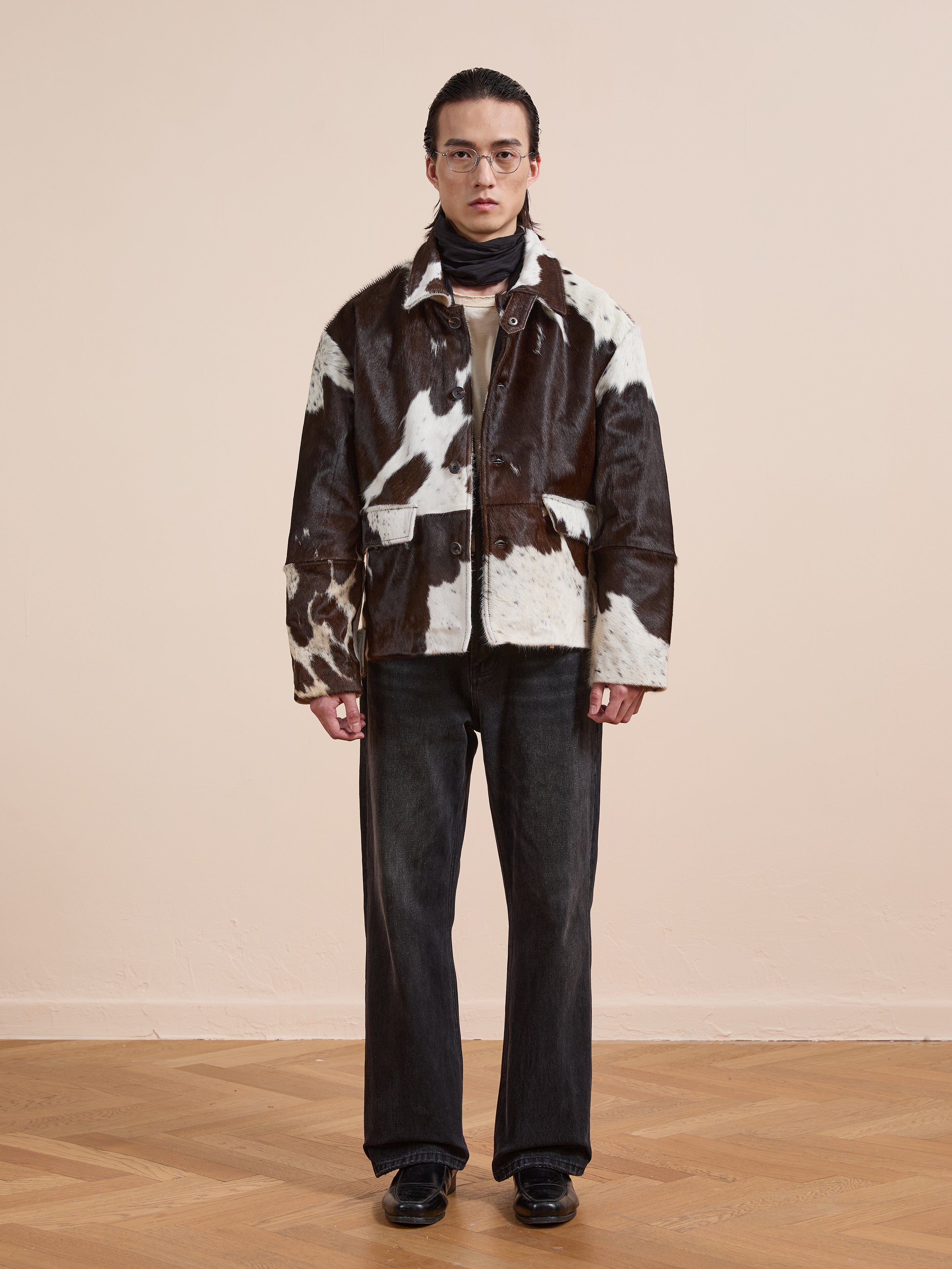 A person stands indoors against a light wall and wooden floor, wearing the FOUND Cowhide Prairie Utility Jacket in brown and white, paired with glasses, dark pants, black shoes, and a scarf.
