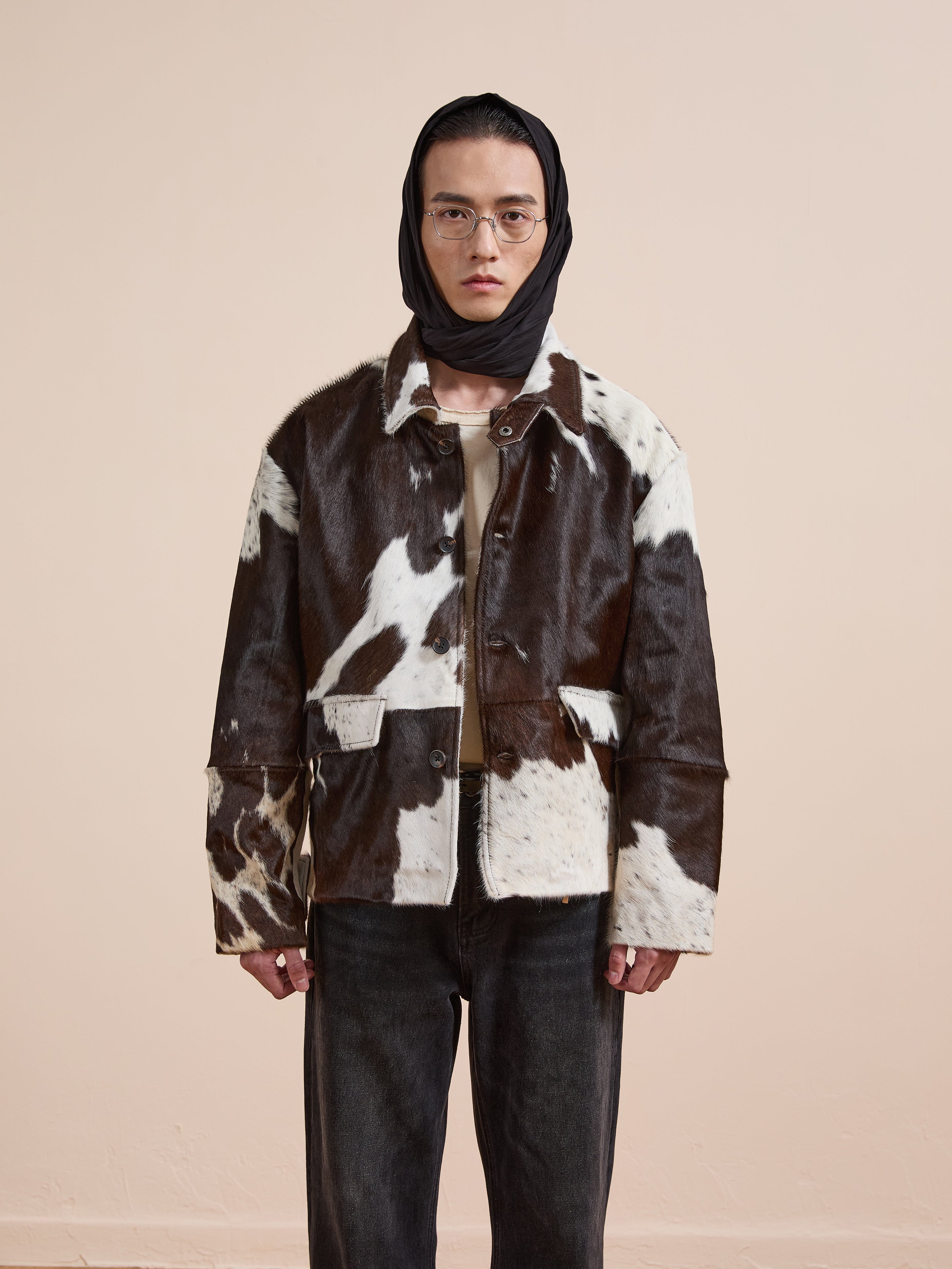 A person faces forward in glasses, a black headscarf, dark jeans, and the FOUND Cowhide Prairie Utility Jacket—brown and white cowhide—set against a plain beige background.