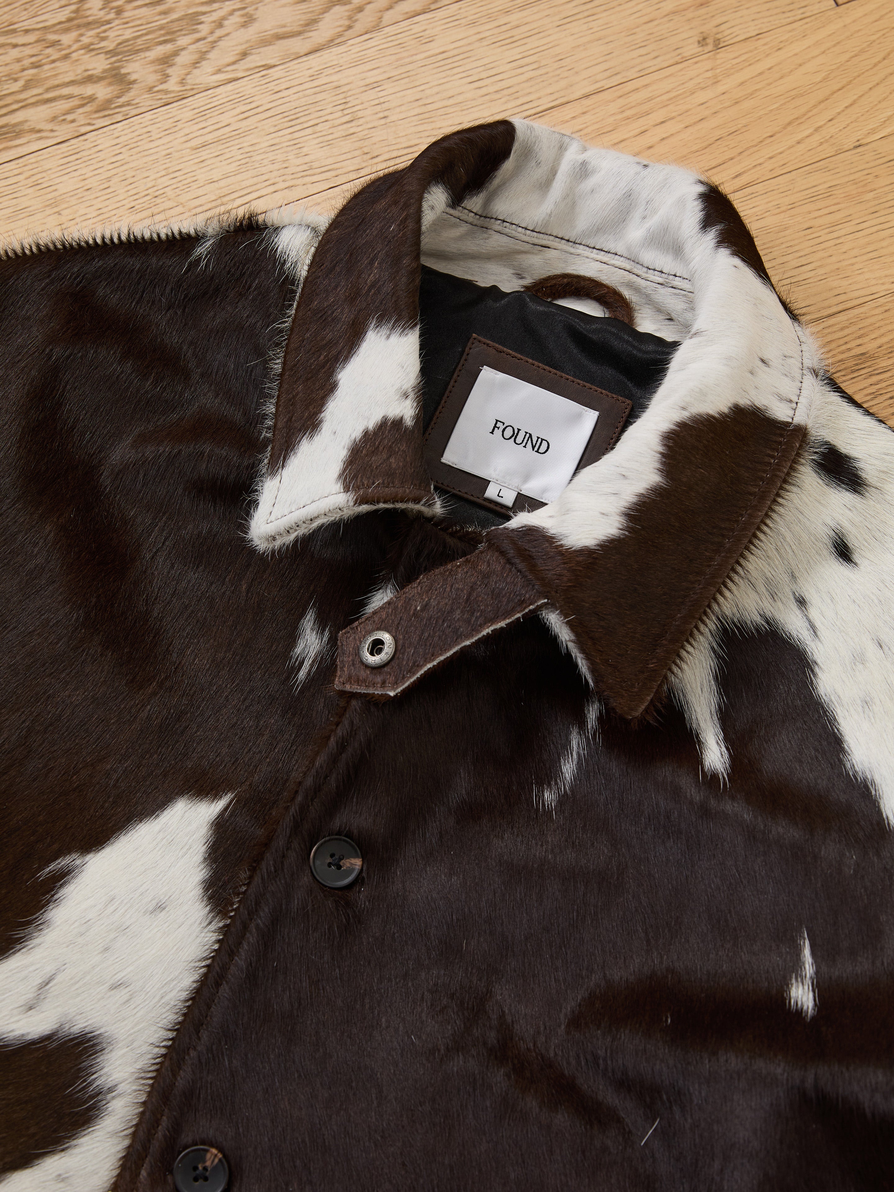 A close-up of the FOUND Cowhide Prairie Utility Jacket, crafted from brown and white cowhide, with the brand label on the inside collar, displayed on a wooden surface.