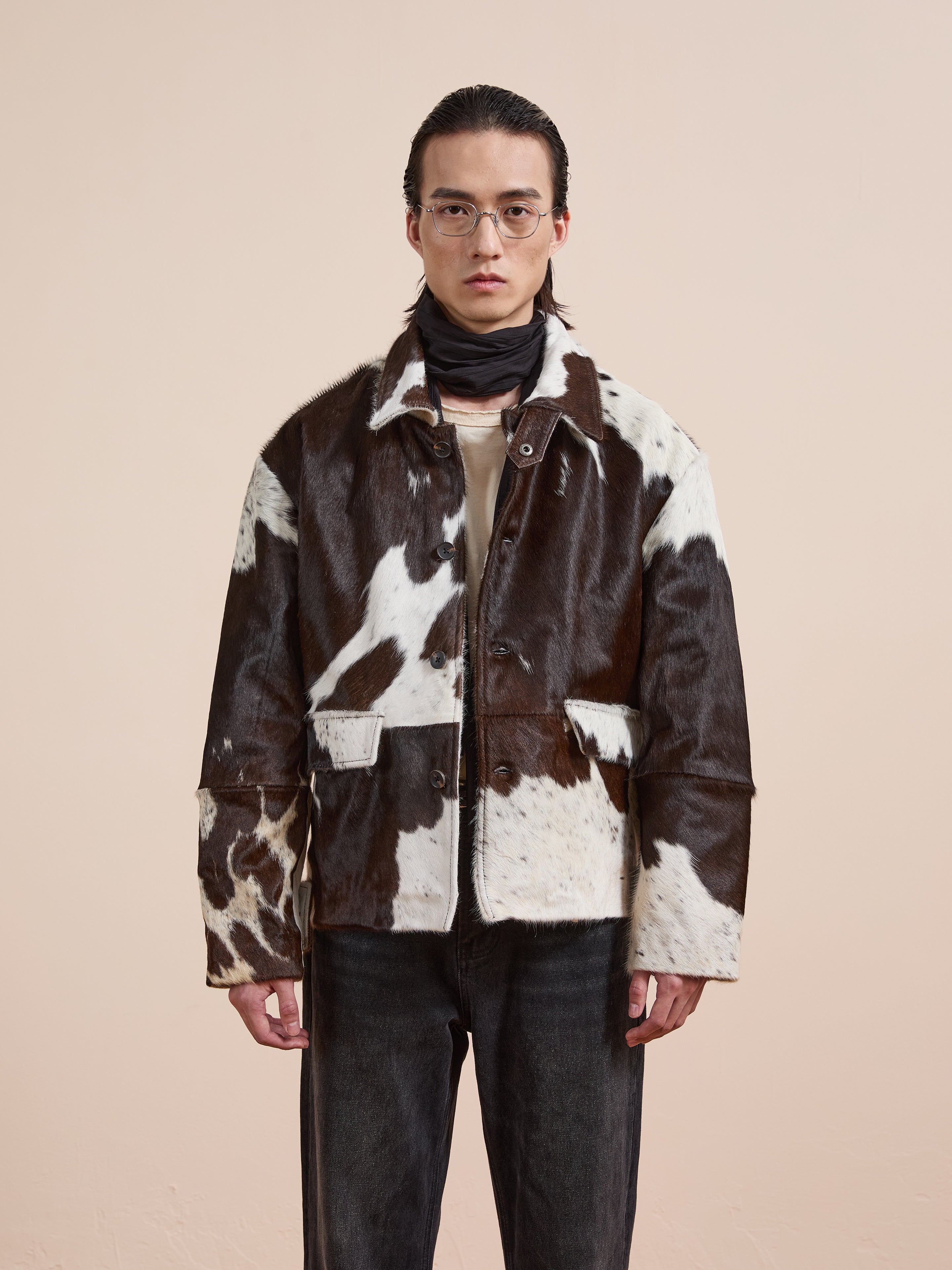 A person wearing glasses, a black and white Cowhide Prairie Utility Jacket by FOUND, a dark scarf, beige shirt, and dark jeans stands facing the camera against a plain light background.