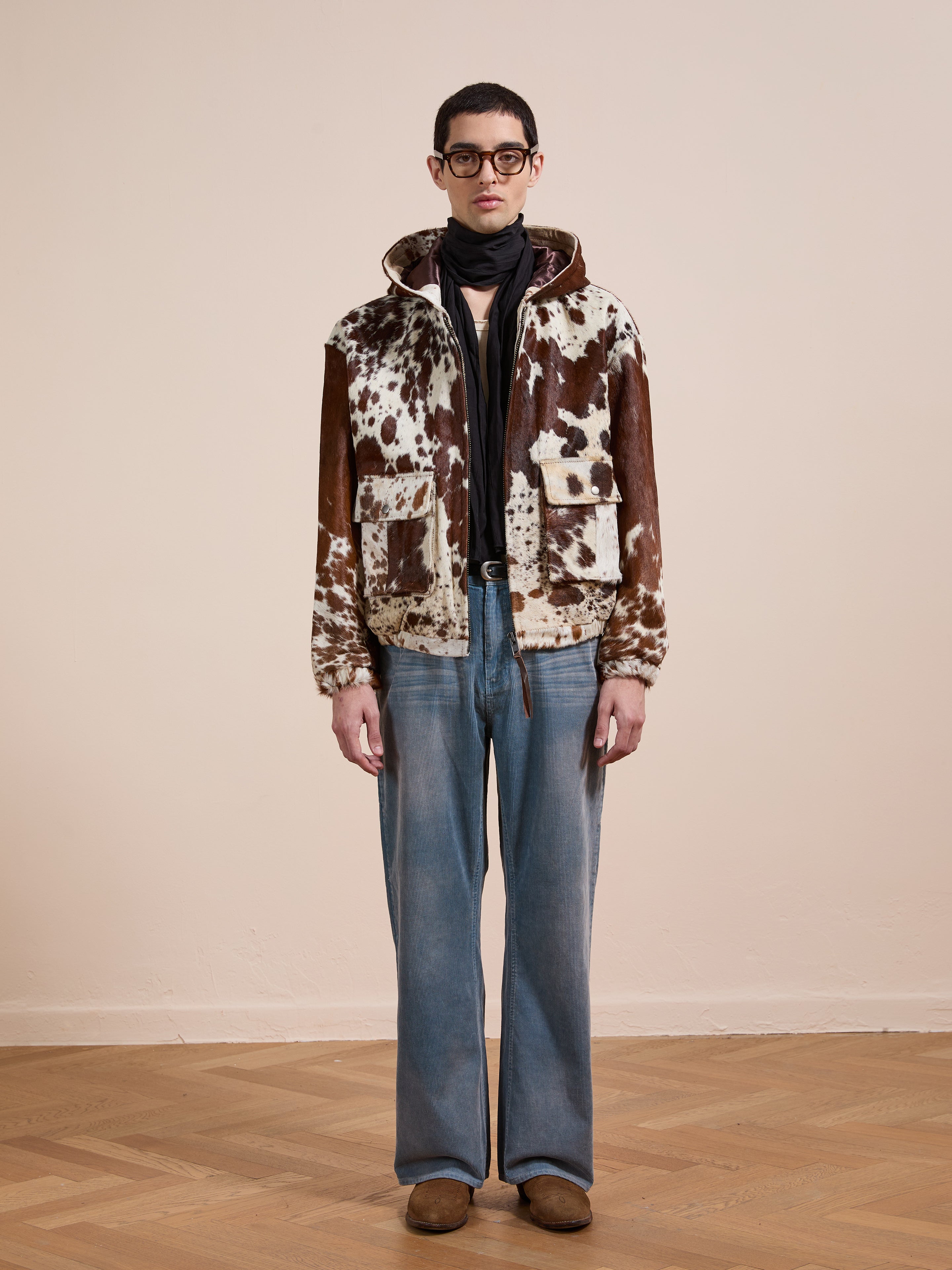 A man in glasses, the FOUND Cowhide Drover Hoodie with brown and white patterns, blue wide-leg jeans, brown shoes, and a black scarf stands indoors against a plain light wall.
