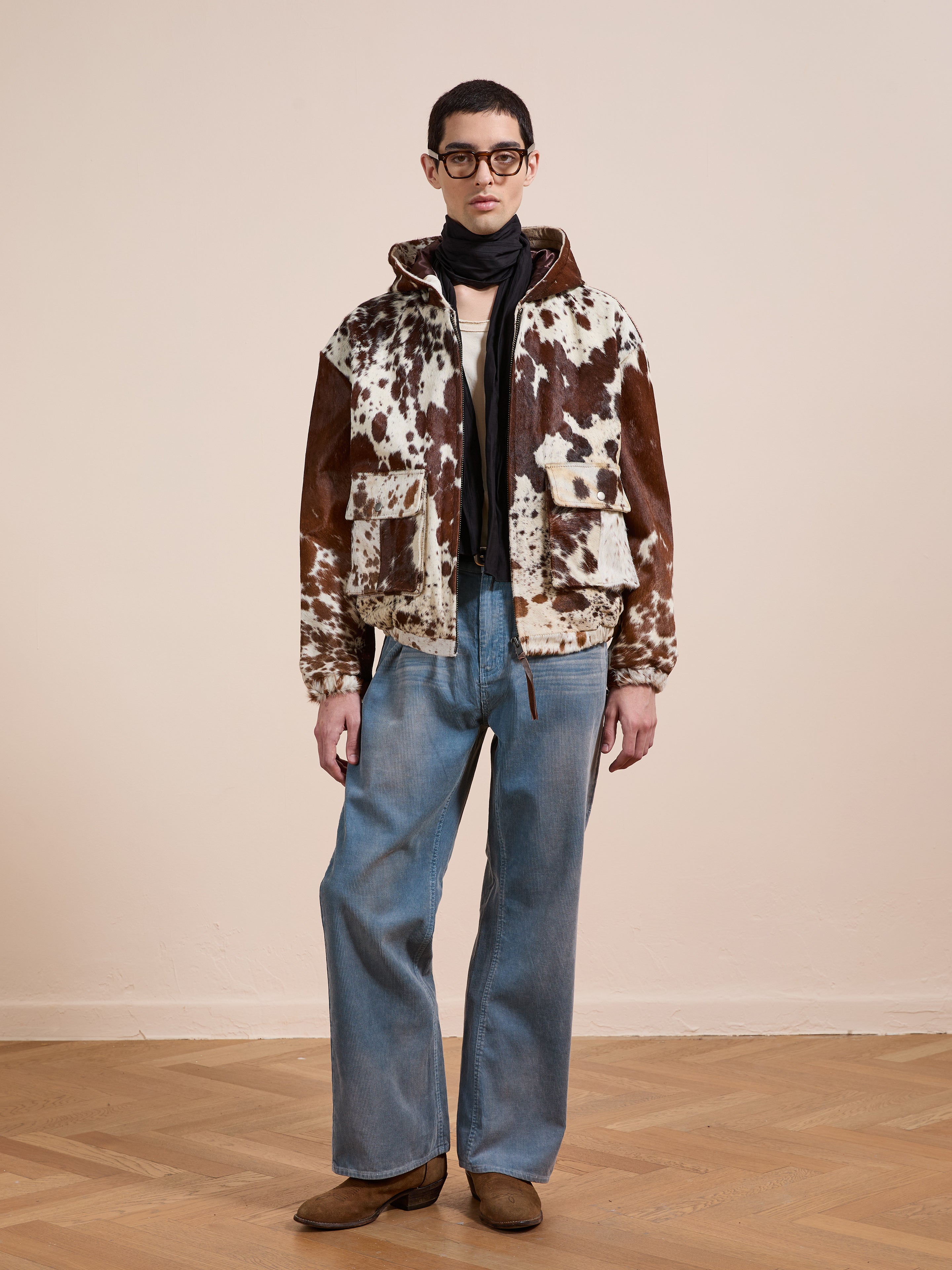 A person stands indoors on a wooden floor against a plain beige wall, wearing FOUND’s Cowhide Drover Hoodie, a black scarf, blue wide-leg jeans, brown boots, and glasses.