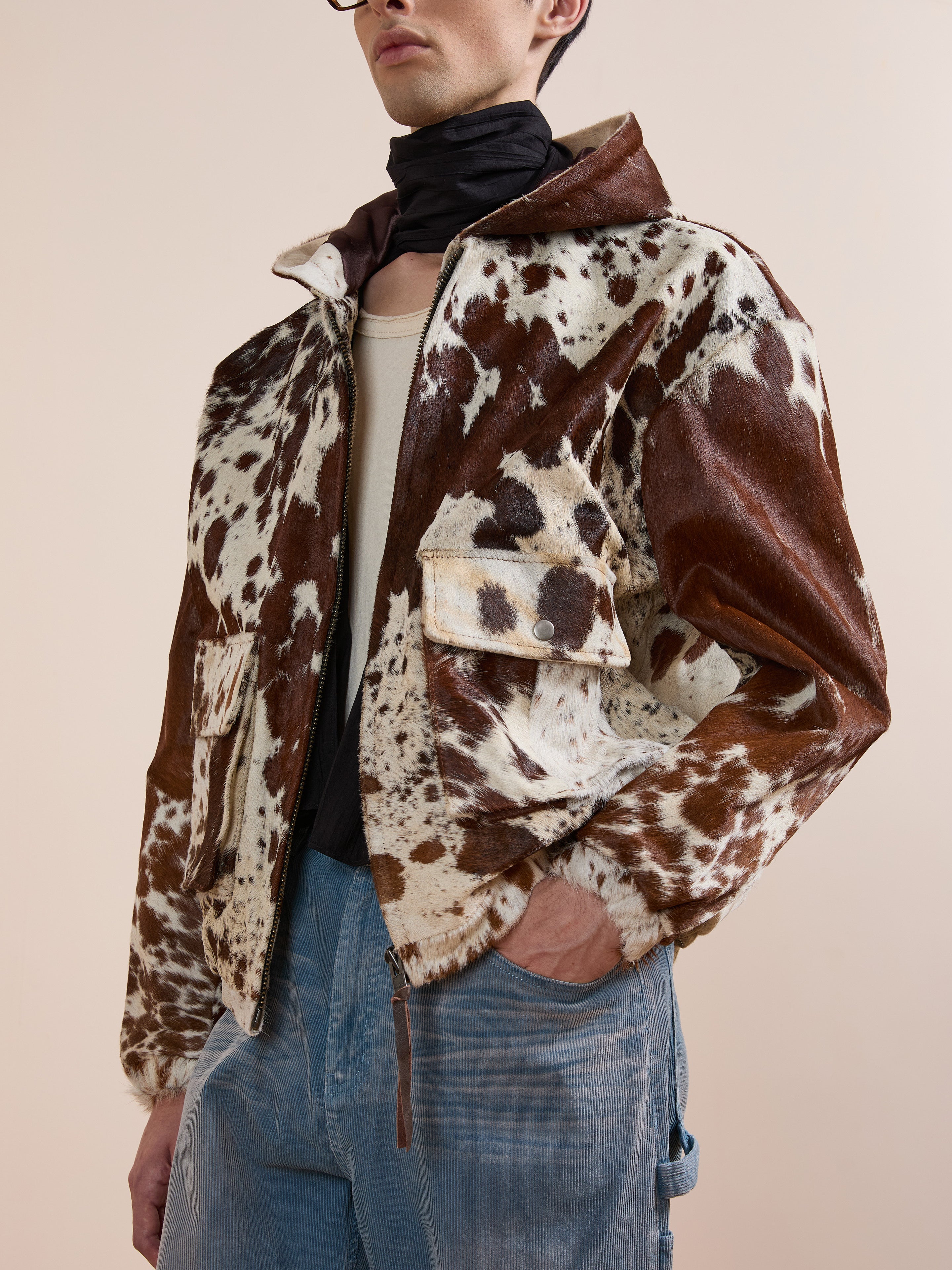 A person wearing the FOUND Cowhide Drover Hoodie in a brown and white pattern, light blue corduroy pants, a beige shirt, and a dark scarf around the neck.