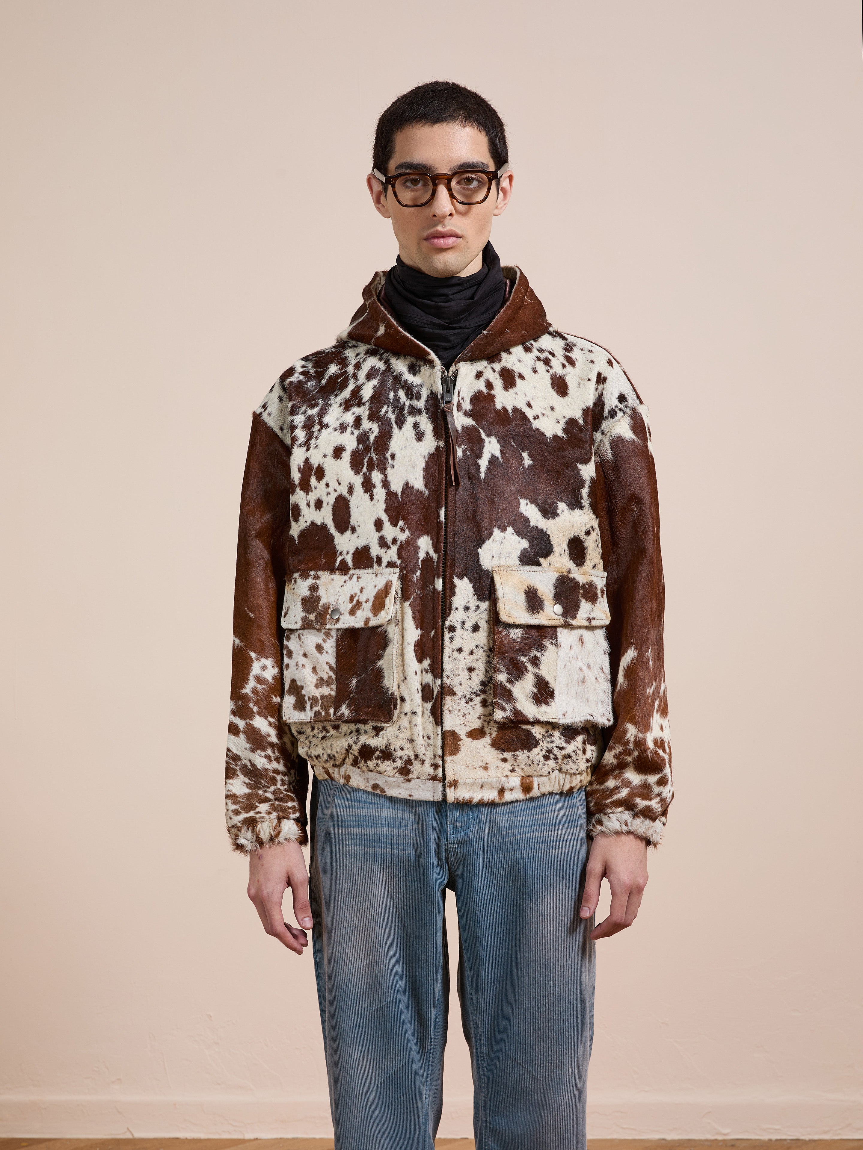 A person in glasses and a black turtleneck wears the FOUND Cowhide Drover Hoodie, featuring a brown and white cow-print design, and stands facing forward against a plain beige background.