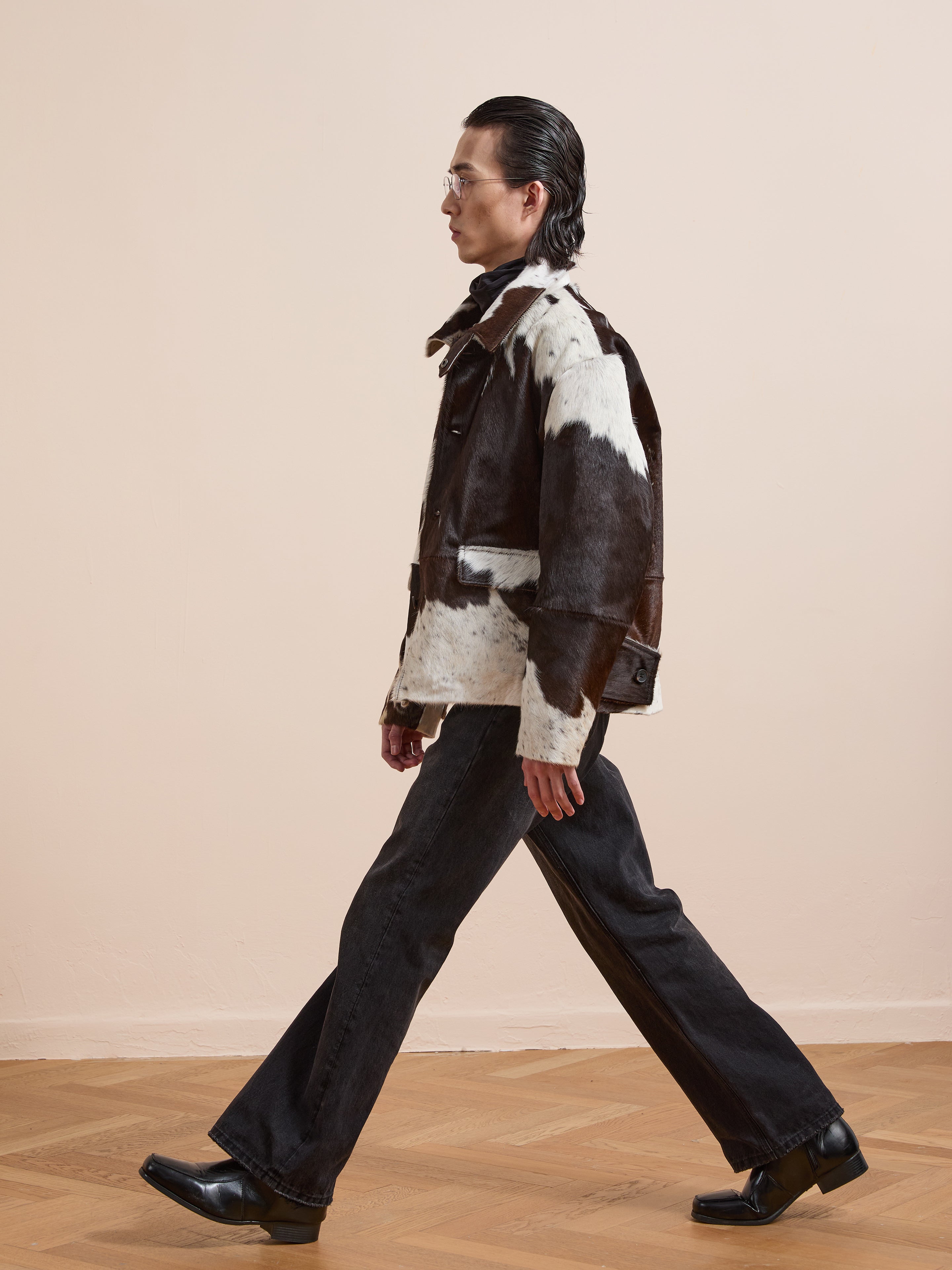 A person with slicked-back hair walks indoors wearing the FOUND Cowhide Prairie Utility Jacket, dark flared jeans, and black square-toe shoes.